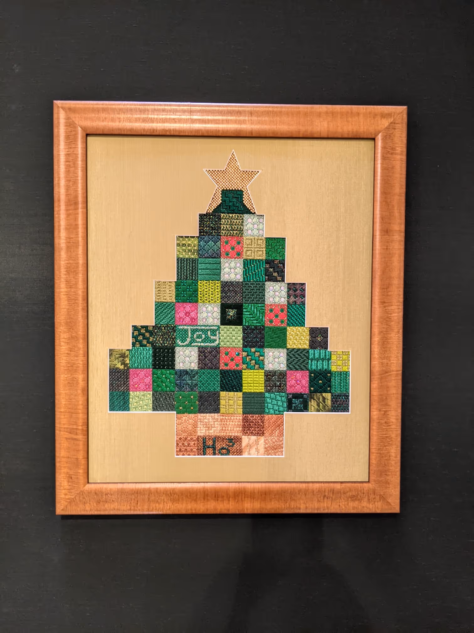 Framed cross-stitch Christmas tree composed of small colorful stitched squares, topped with a stitched star and words 'Joy' and 'Ho' in the design.