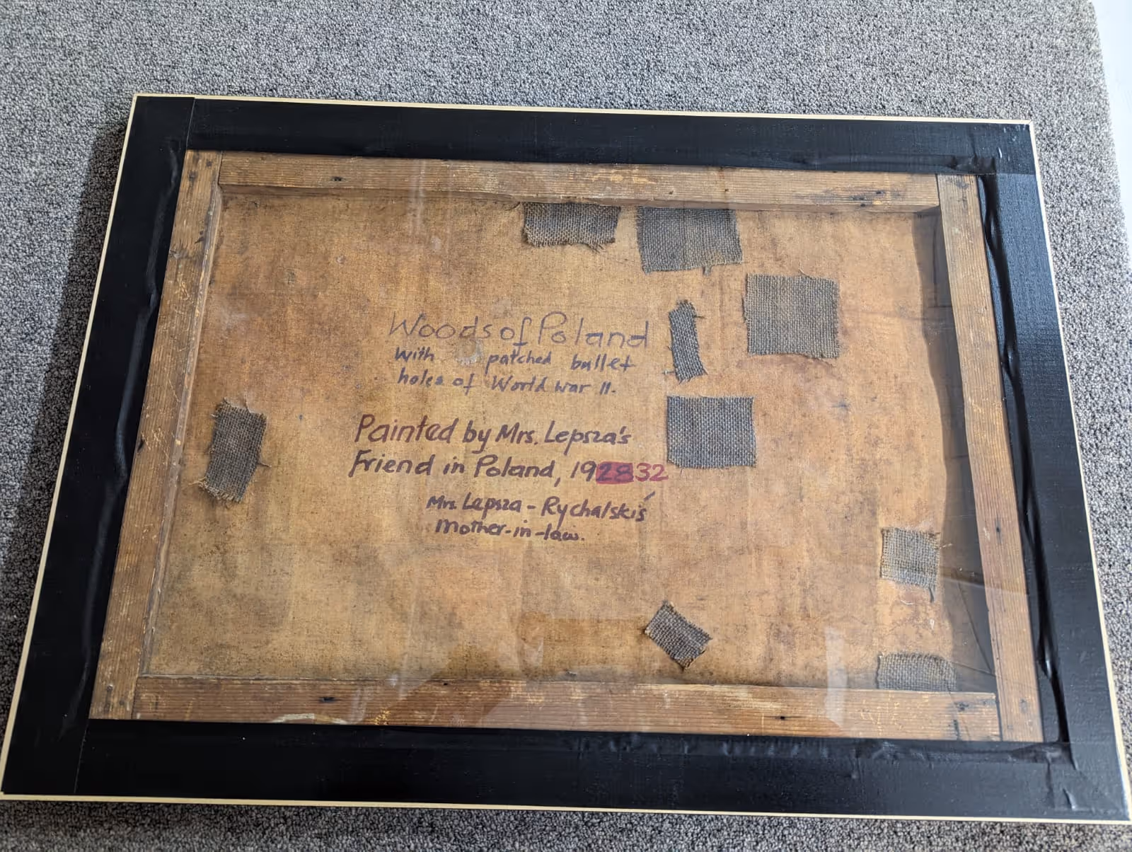 Back of a framed canvas with handwritten text describing it as 'Woods of Poland' with patched bullet holes from World War II, painted by Mrs. Lepza's friend in Poland in 1932, including several fabric patches.