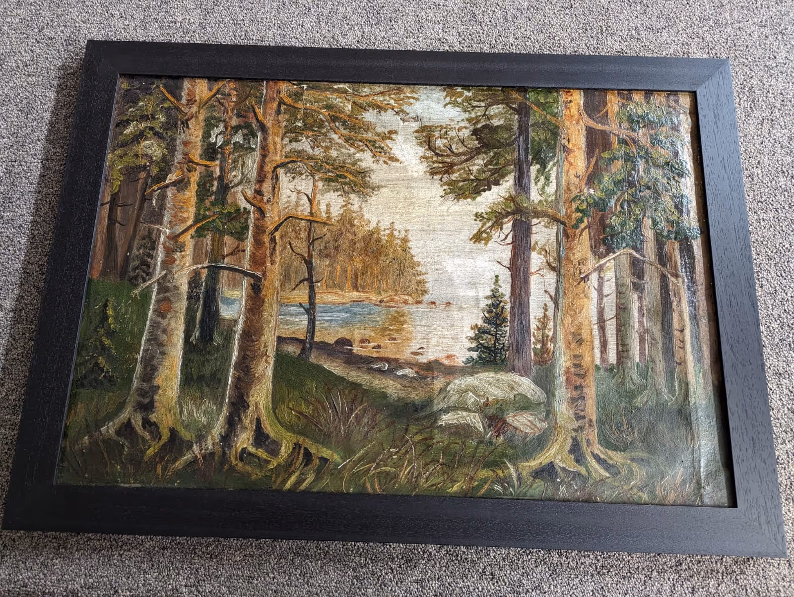 Framed painting of a forest with tall trees, rocks, grassy foreground, and a river or lake in the background.