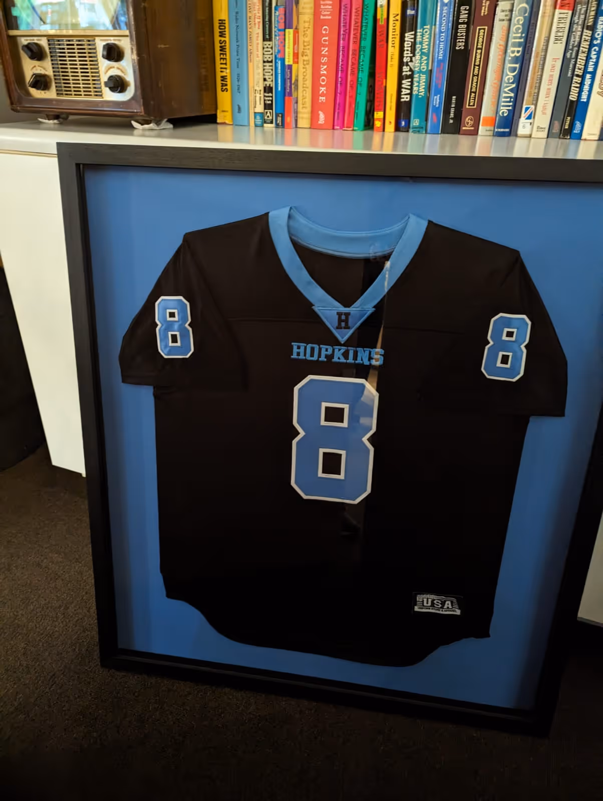 Framed black Hopkins sports jersey with light blue accents and the number 8 on the front and sleeves, displayed against a blue background.