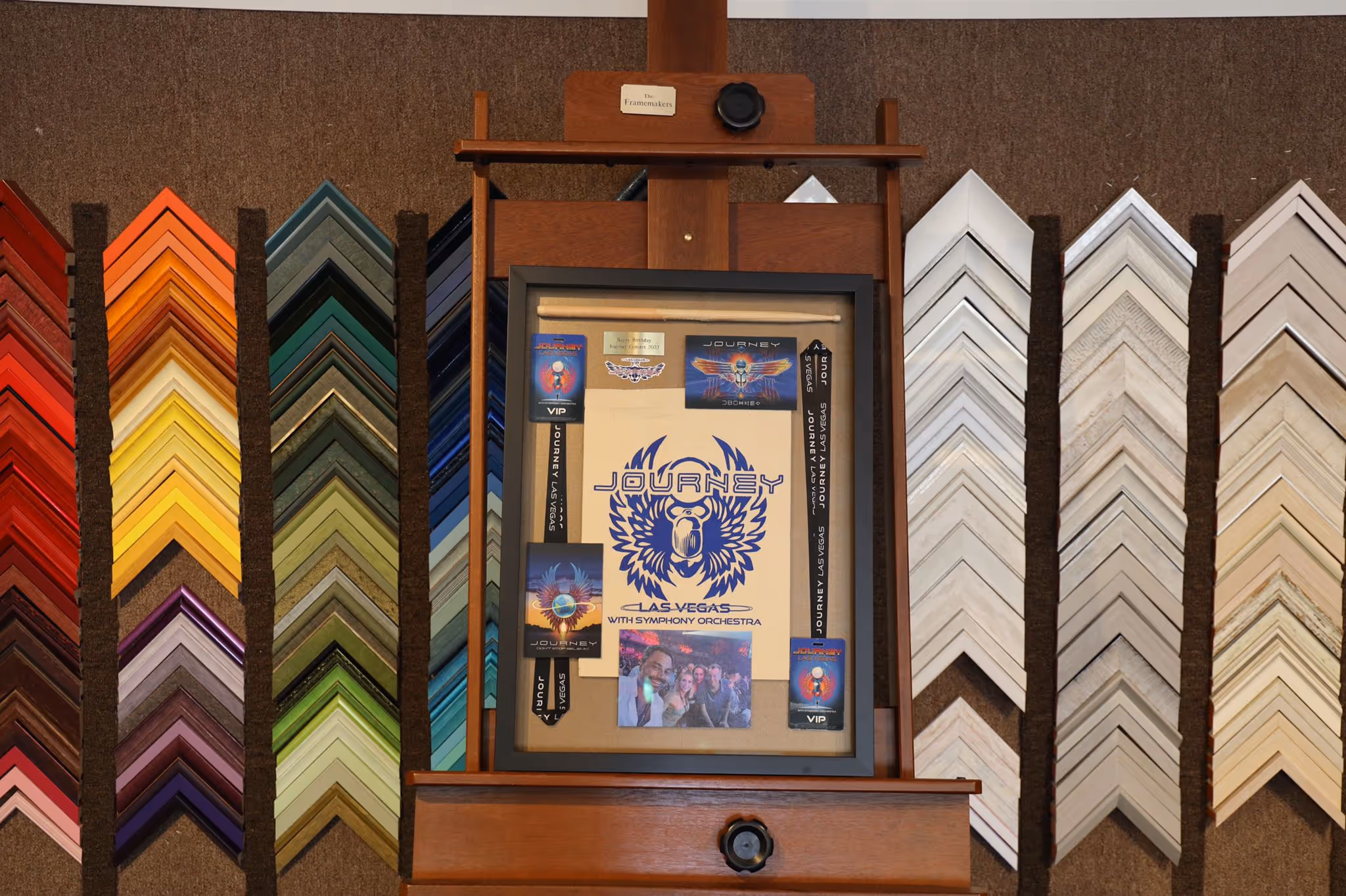 An easel with a framed collage featuring Journey Las Vegas concert memorabilia surrounded by various colorful picture frame corner samples on the wall.