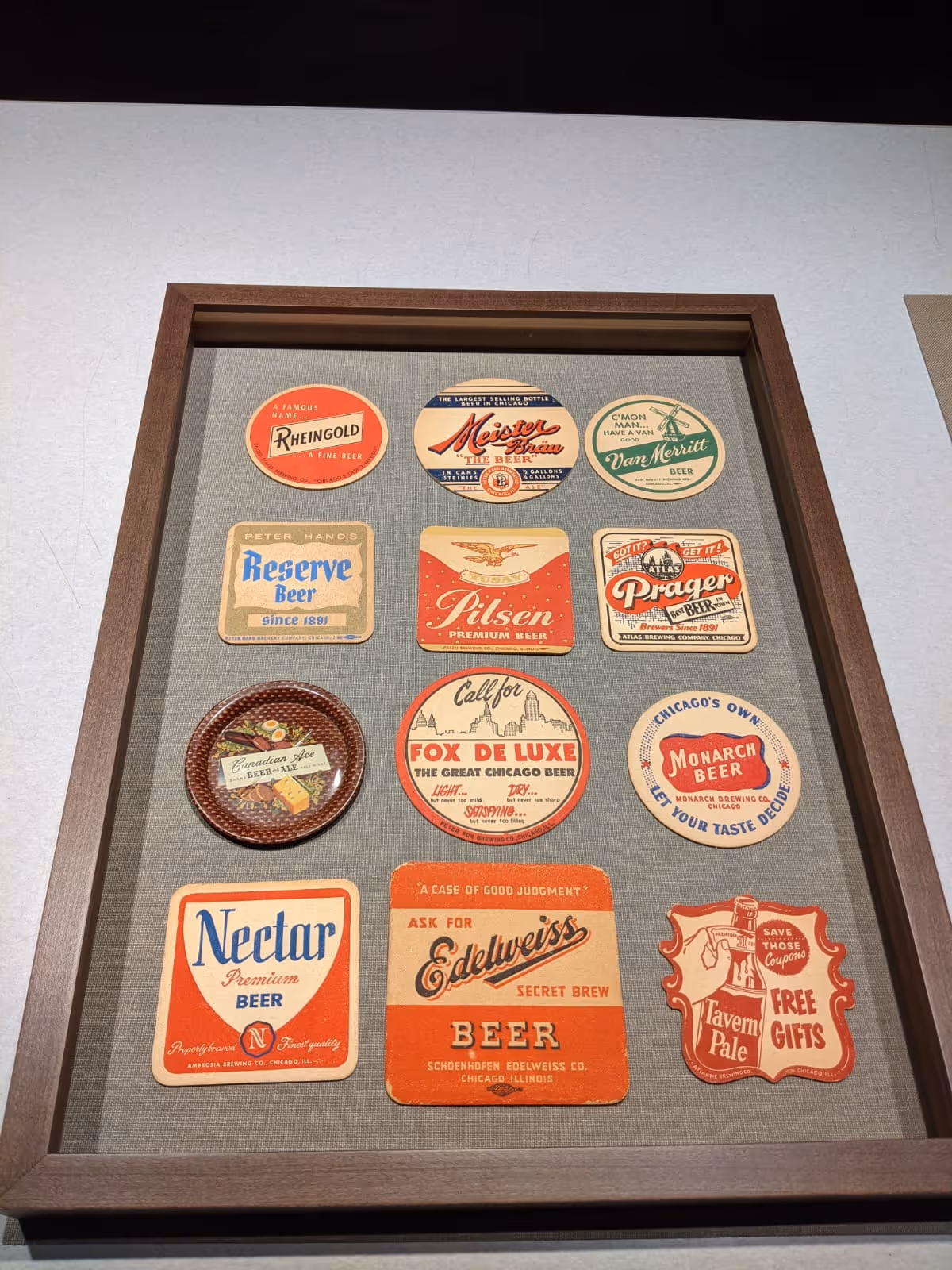 Framed display of twelve vintage beer coasters and labels arranged in three rows on a grey fabric background.