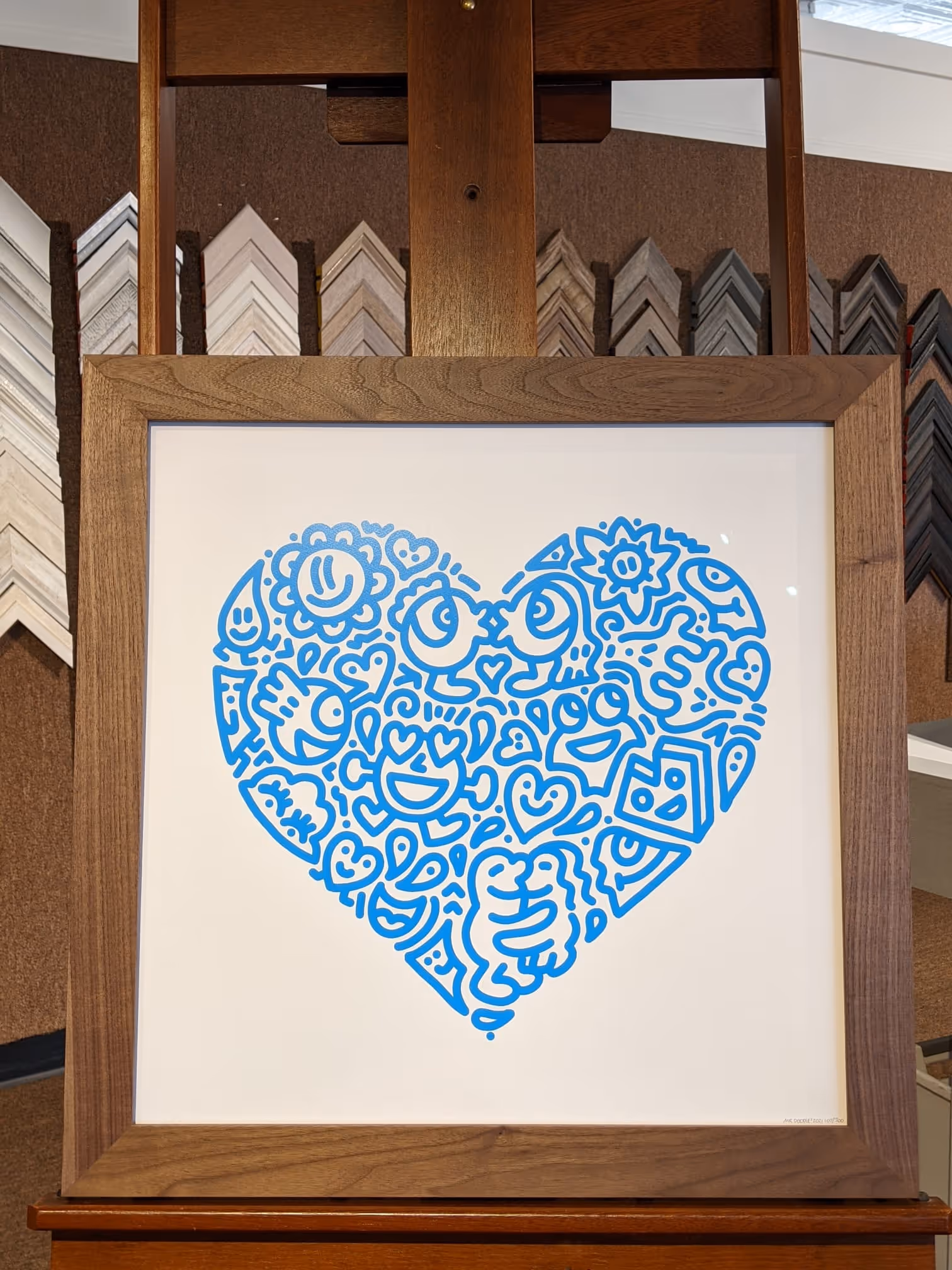 Framed artwork of a large blue heart composed of abstract doodles including faces, hearts, and flowers.