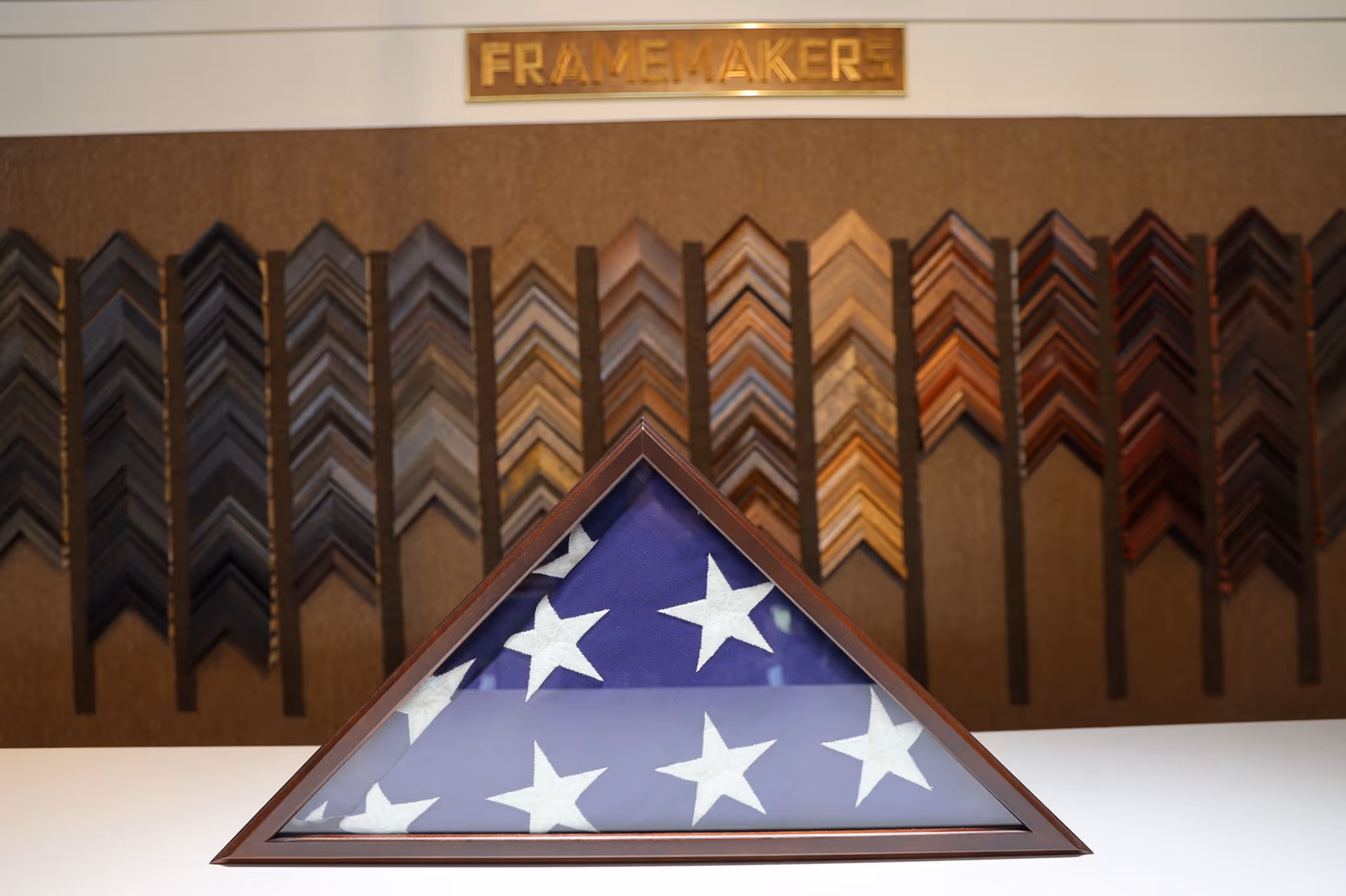 Folded American flag with white stars in a triangular wooden display case on a white surface, with various wooden frame samples on the wall behind it under a sign reading 'FRAMEMAKERS'.