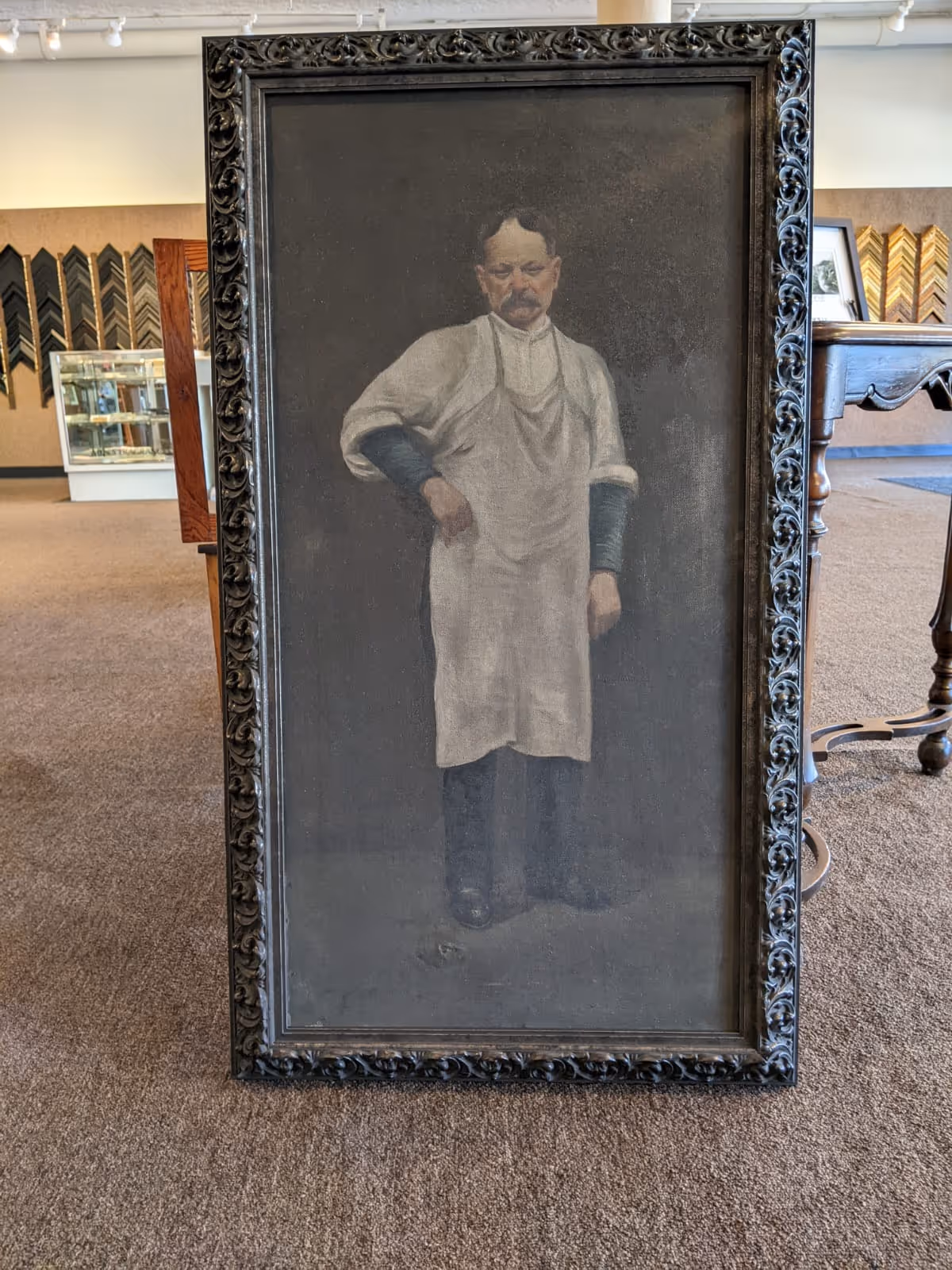 Framed painting of a man with a mustache wearing a white apron and dark sleeves, standing with one hand on his hip.