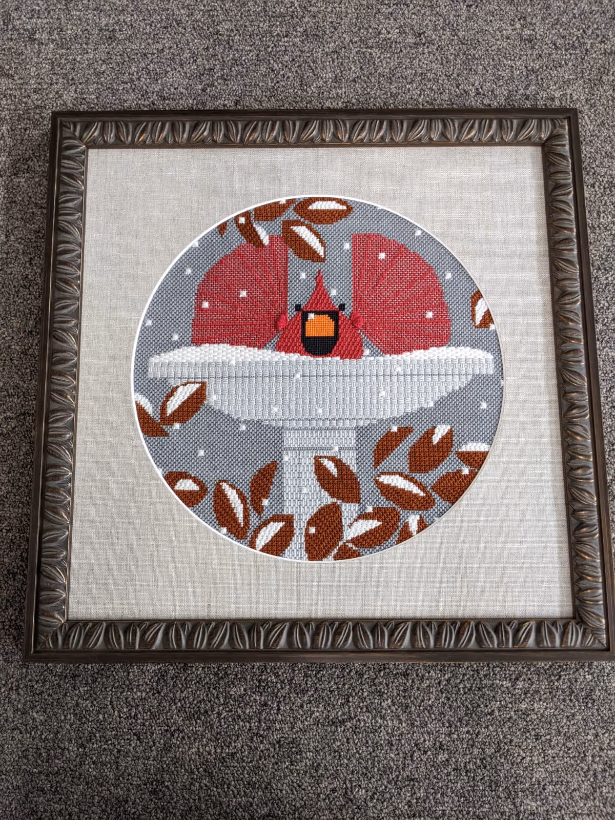 Framed cross-stitch art of a red cardinal perched on a snow-covered birdbath surrounded by brown leaves with white snowflakes falling on a gray background.