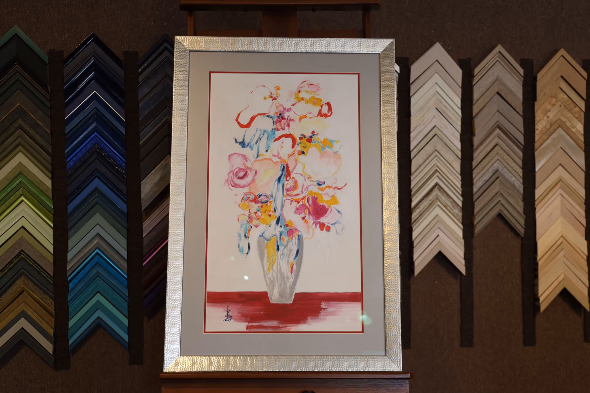 Abstract floral painting in a silver frame displayed against a wall with various colorful picture frame samples.