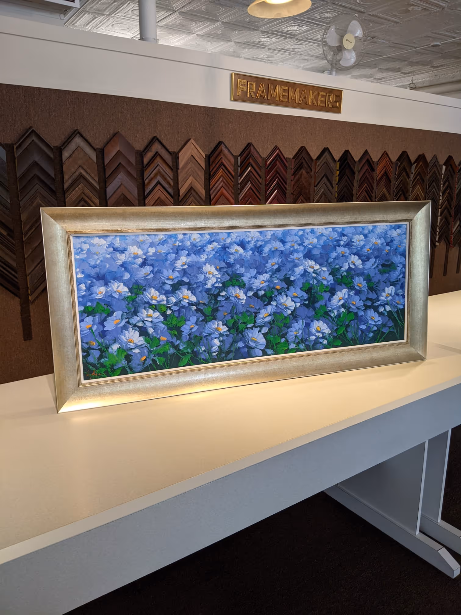 Framed painting of dense blue and white flowers on a white table in a frame shop.
