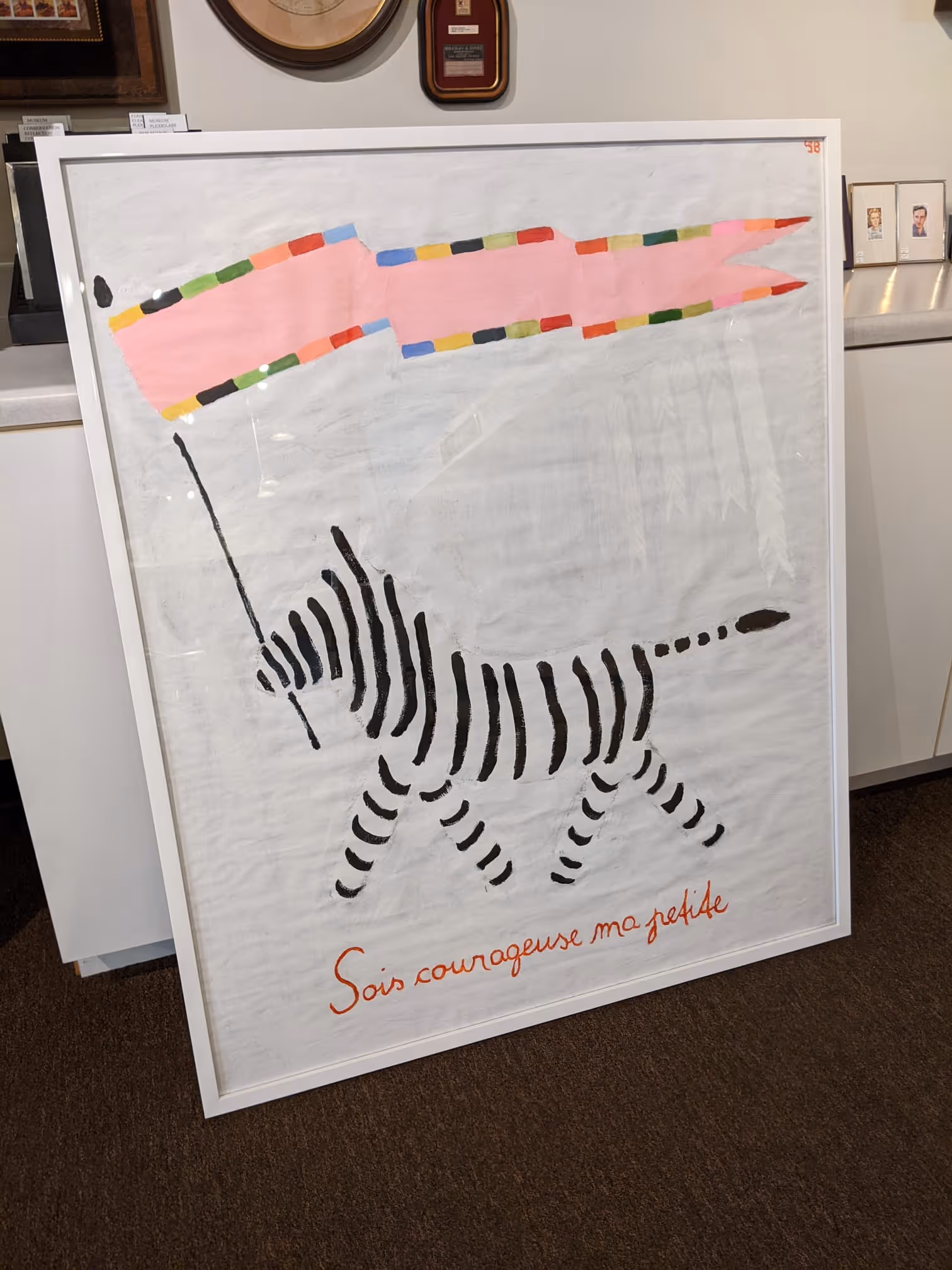 Framed artwork of a stylized black and white striped zebra holding a colorful, rectangular kite, with red handwritten French text below.