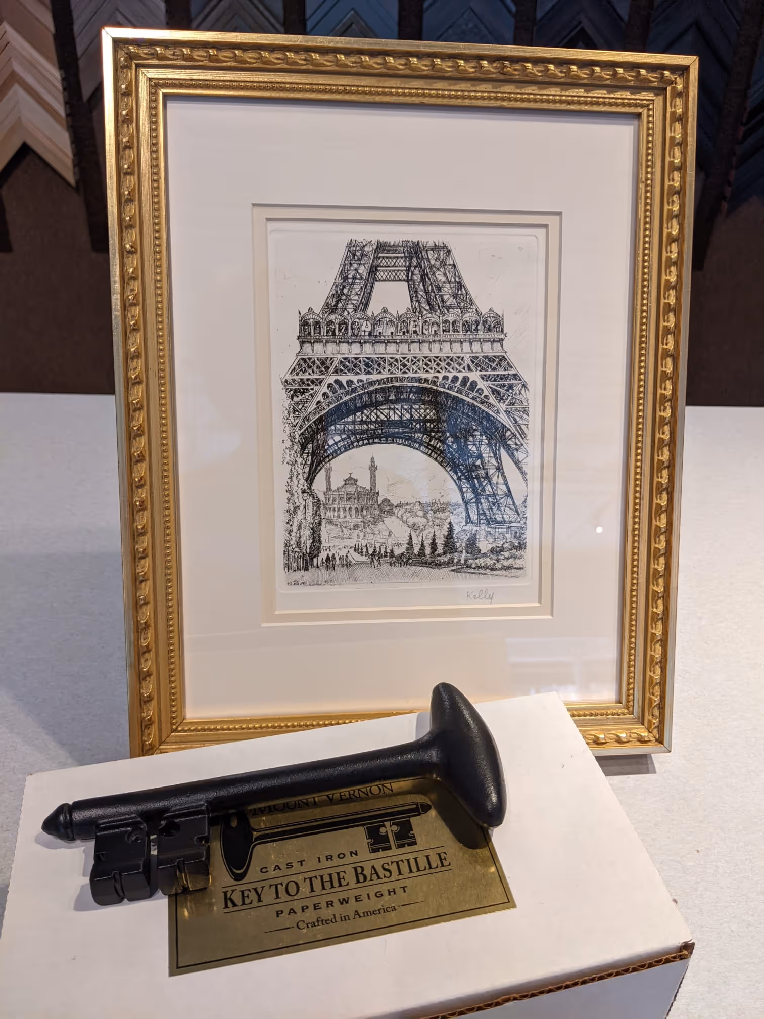 Black cast iron paperweight shaped like a large key labeled 'Key to the Bastille' resting on a white box, with a framed sketch of the Eiffel Tower in the background.