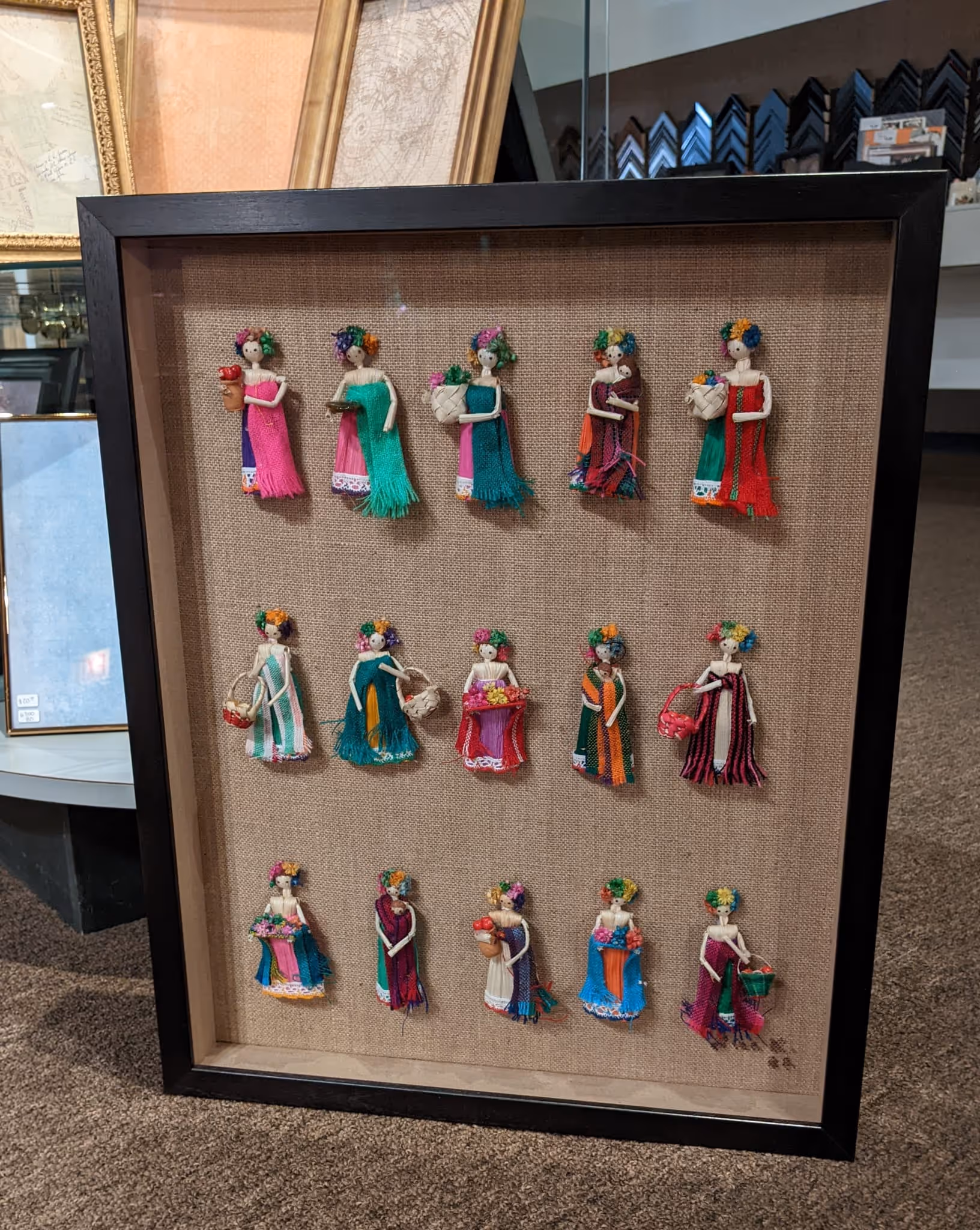 Framed display of fifteen small handcrafted dolls wearing colorful woven dresses and carrying miniature baskets or flowers.