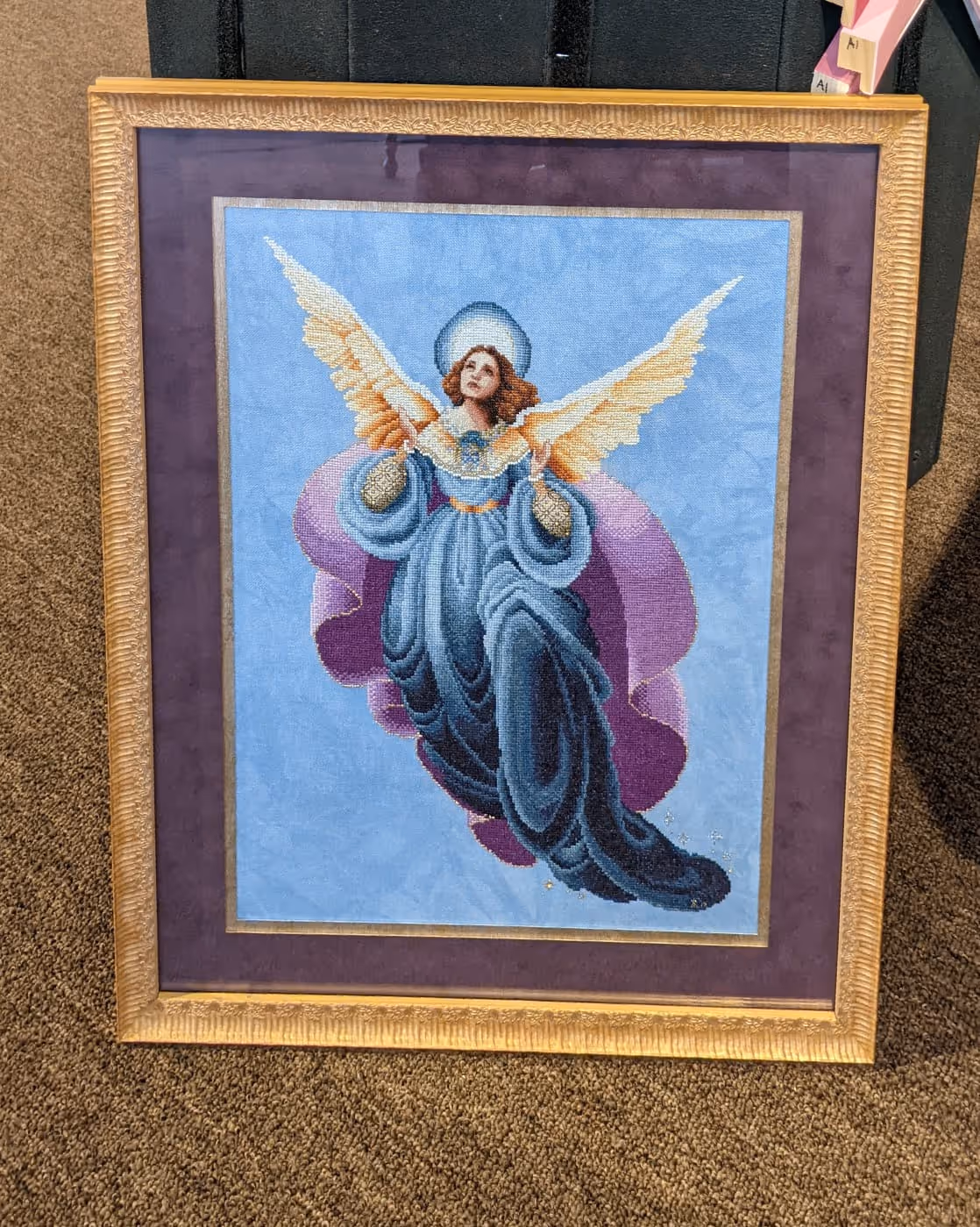 Framed cross-stitch artwork of an angel with golden wings, a blue flowing gown, and a violet background.