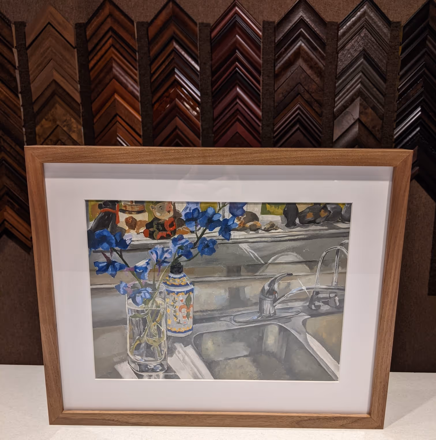 Framed painting of a kitchen sink with a glass vase holding blue flowers and a painted bottle on the counter, with a reflective backsplash.