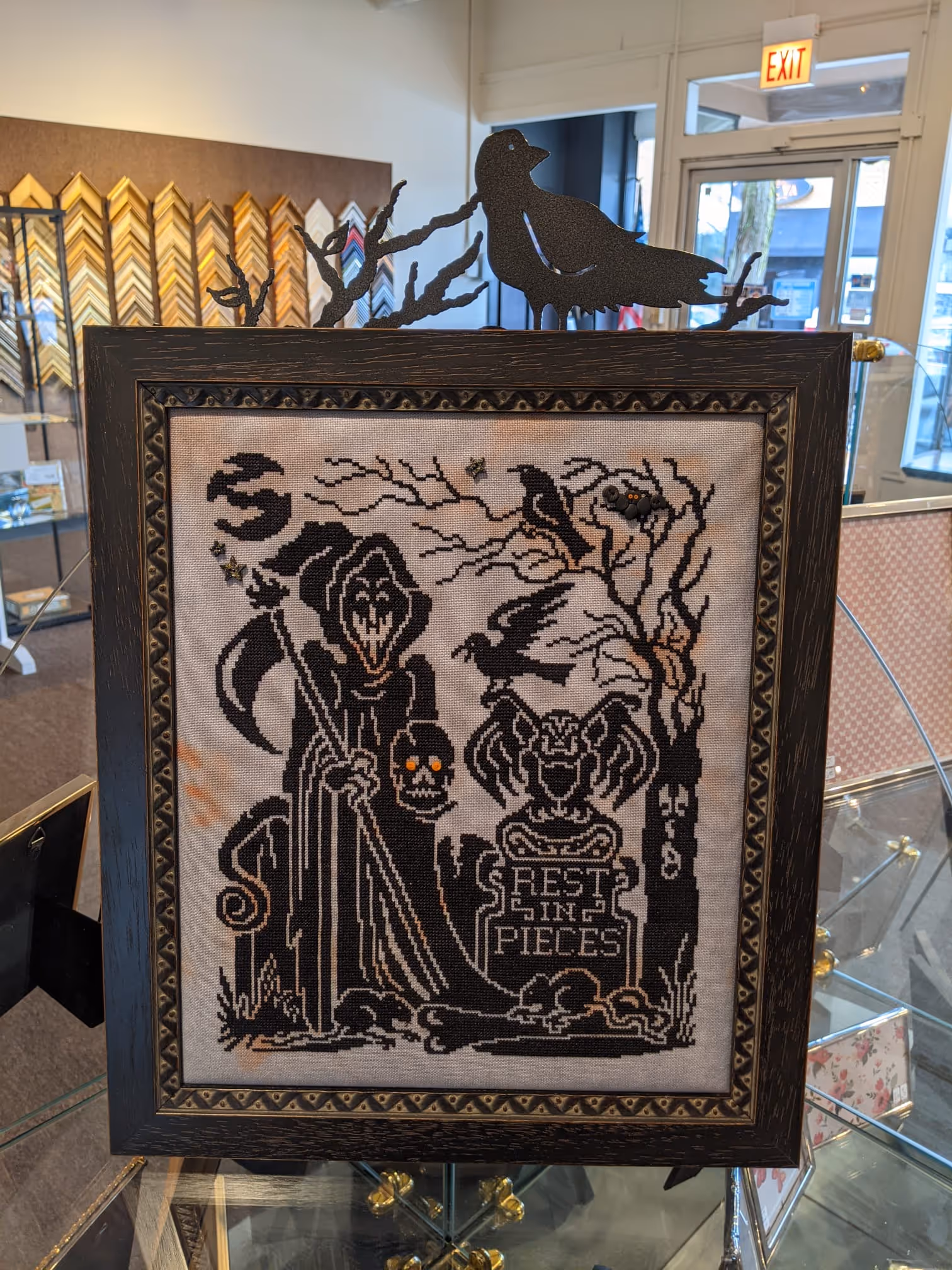 Framed black and white cross-stitch art featuring a grim reaper holding a scythe and a skull with glowing eyes, a tombstone with 'Rest In Pieces', bats, a crow on a tree, and a black crow decoration on top.