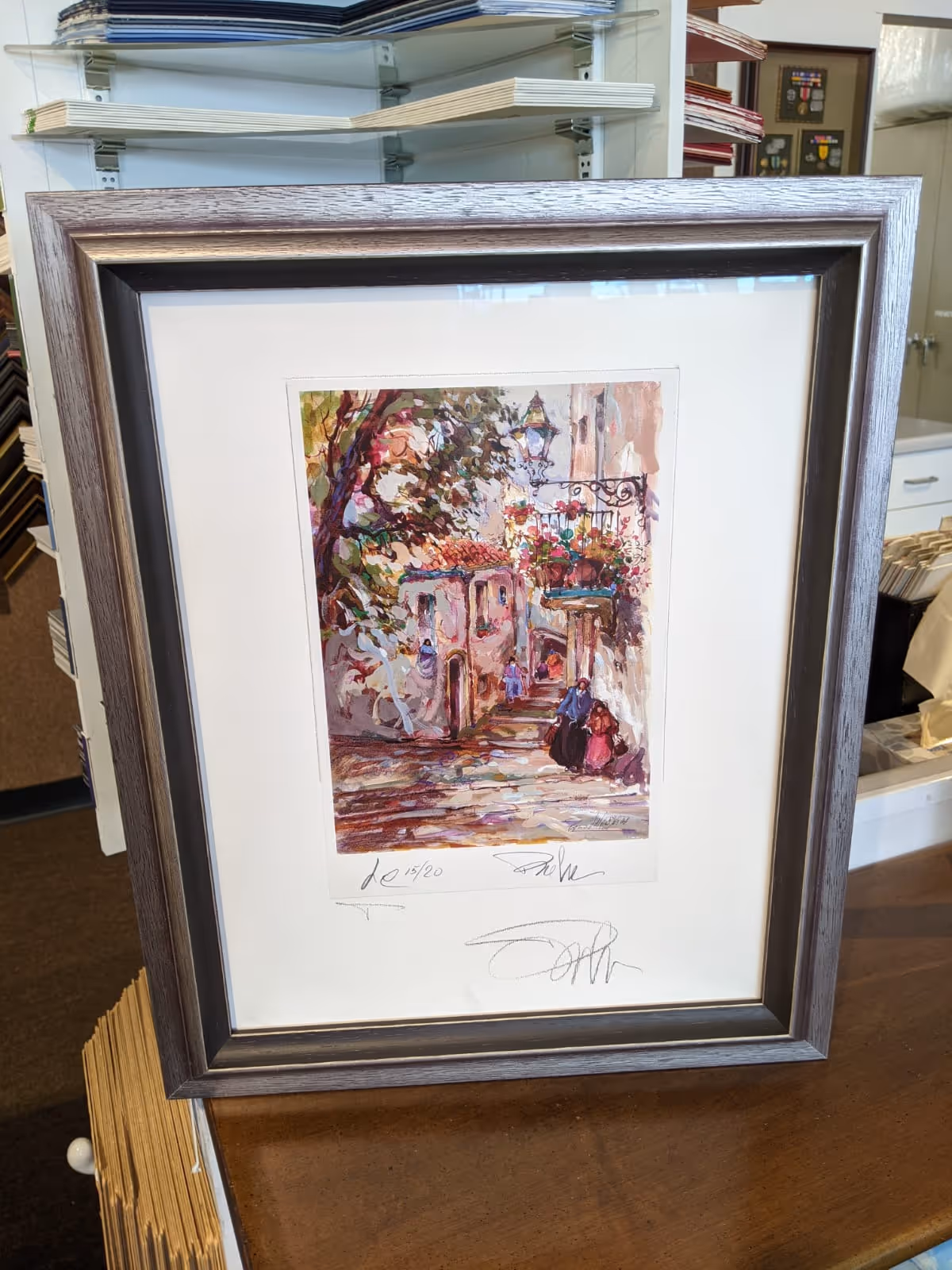 Framed painting of a colorful street scene with two people walking and a balcony adorned with flowers.