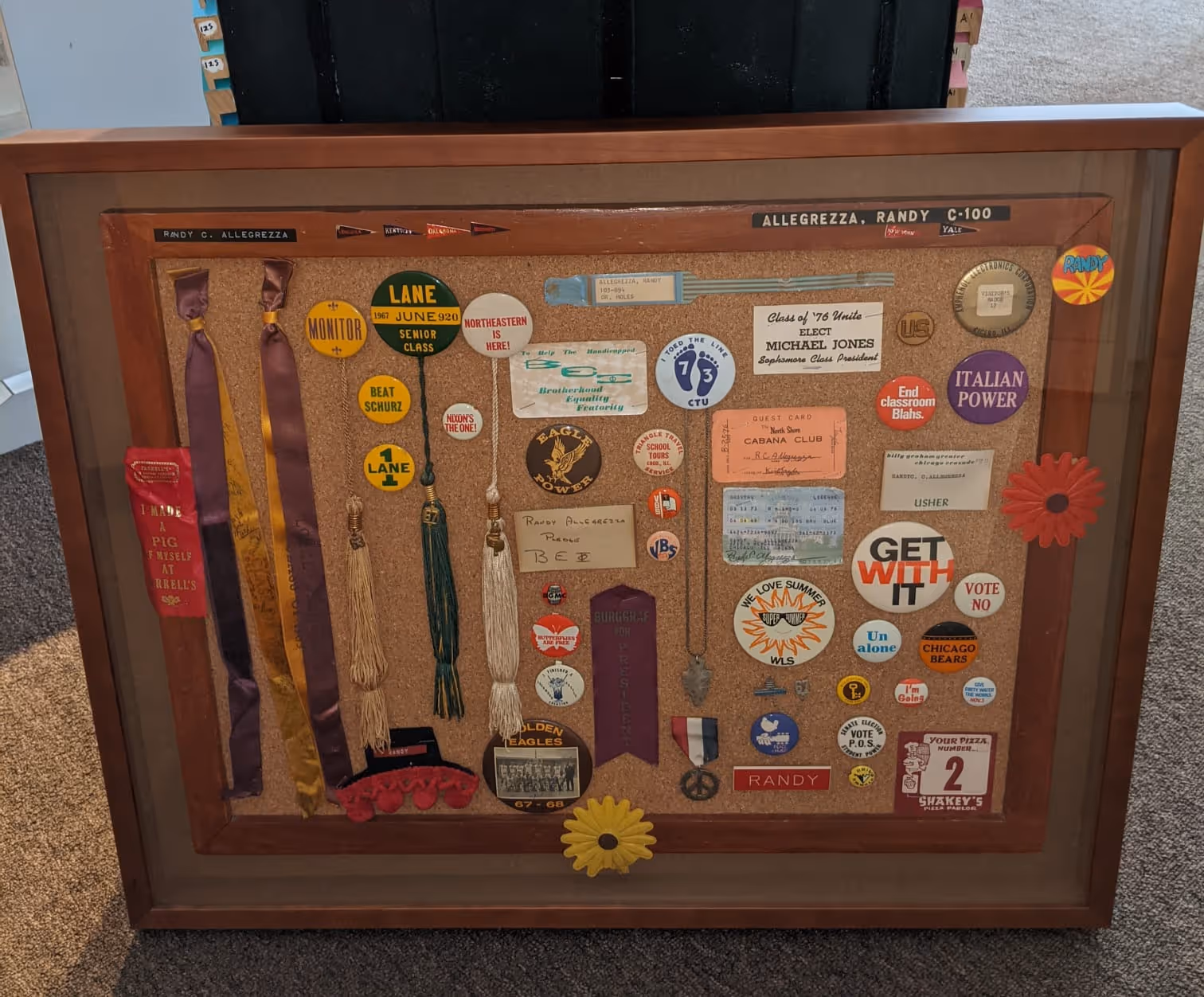 Framed corkboard display with various vintage pins, ribbons, tassels, badges, and memorabilia from Randy C. Allegrezza's school and scouting achievements.