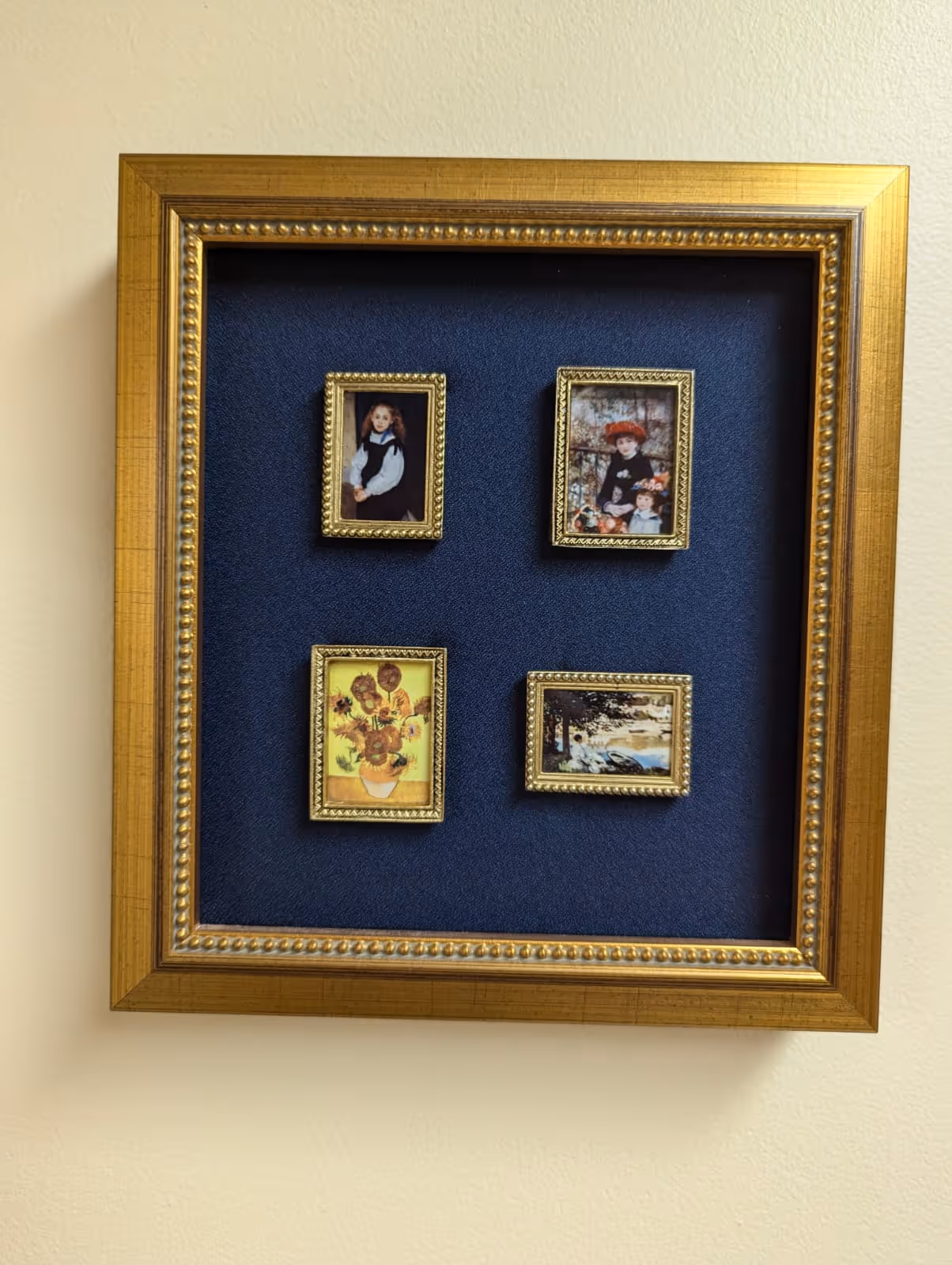 Four miniature framed paintings mounted on a navy blue background inside a larger gold frame on a beige wall.