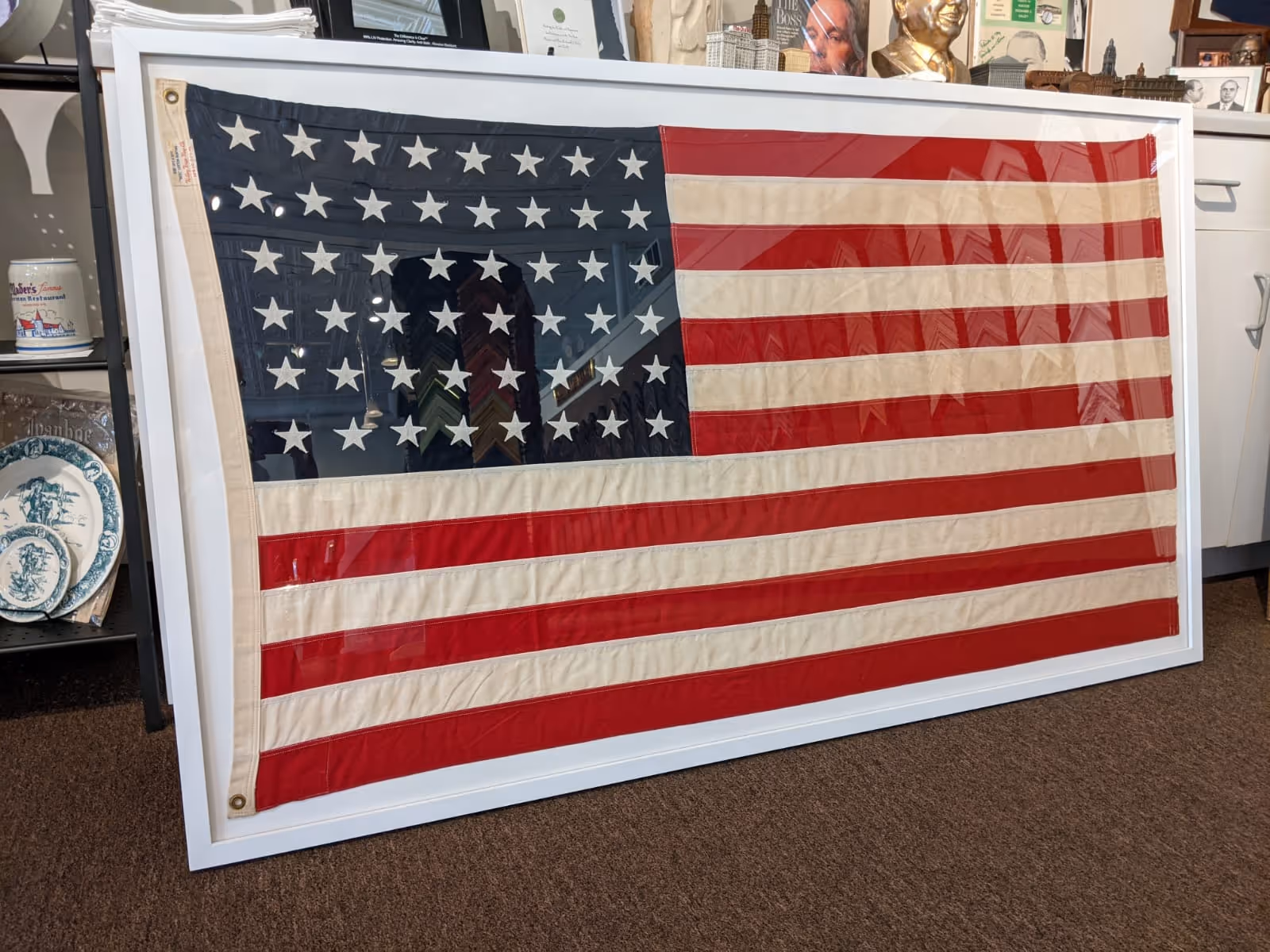 Framed vintage American flag with 48 stars displayed leaning against a cabinet in a room with various decorative items.