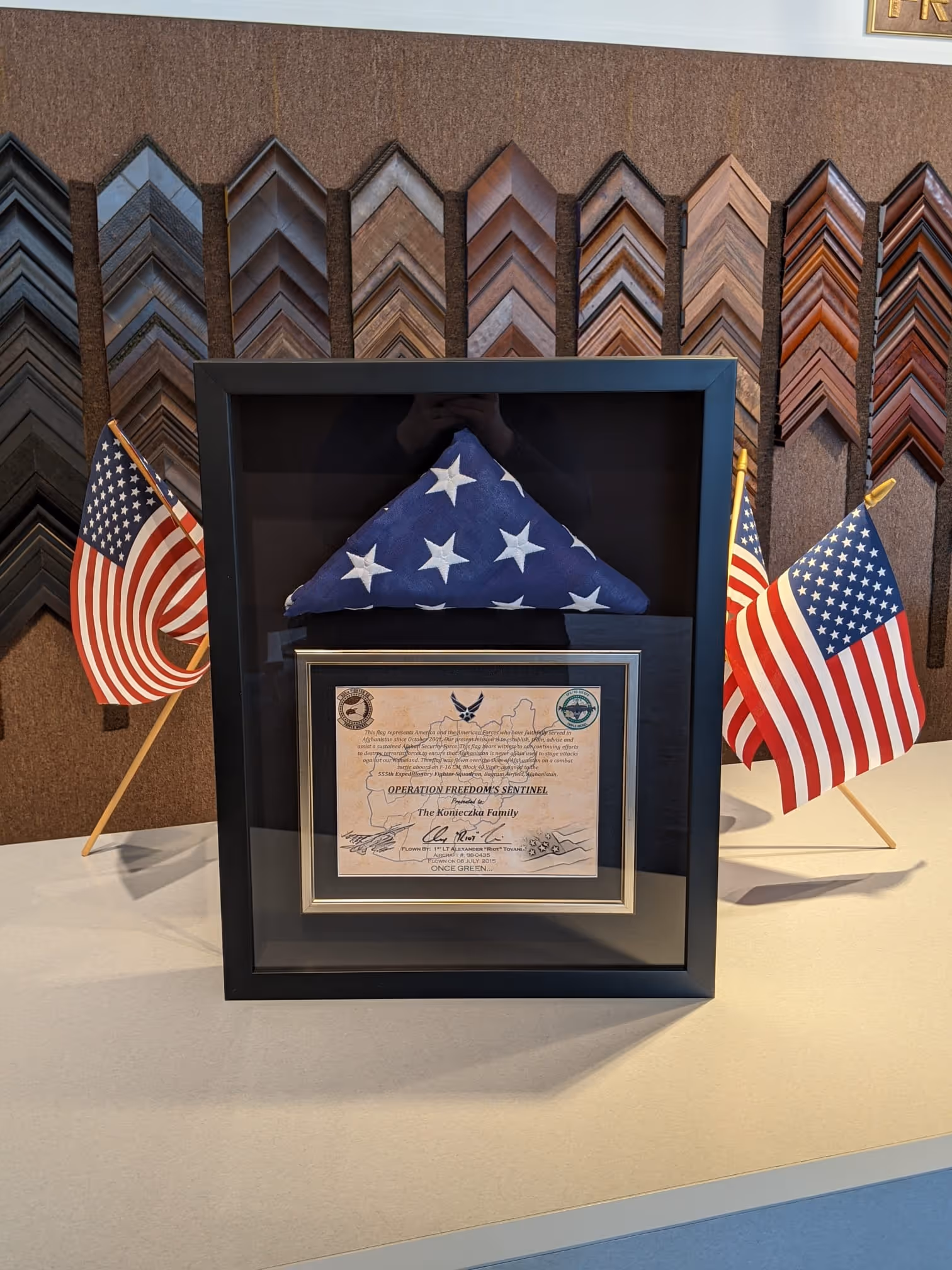 Framed military honor display with a folded American flag and a certificate presented to the Konieczka Family, flanked by two small American flags on a white surface.