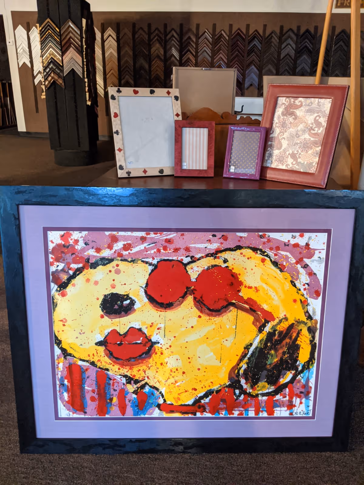Abstract colorful painting of a yellow face with red lips and sunglasses, framed in black and purple, displayed with other empty picture frames.
