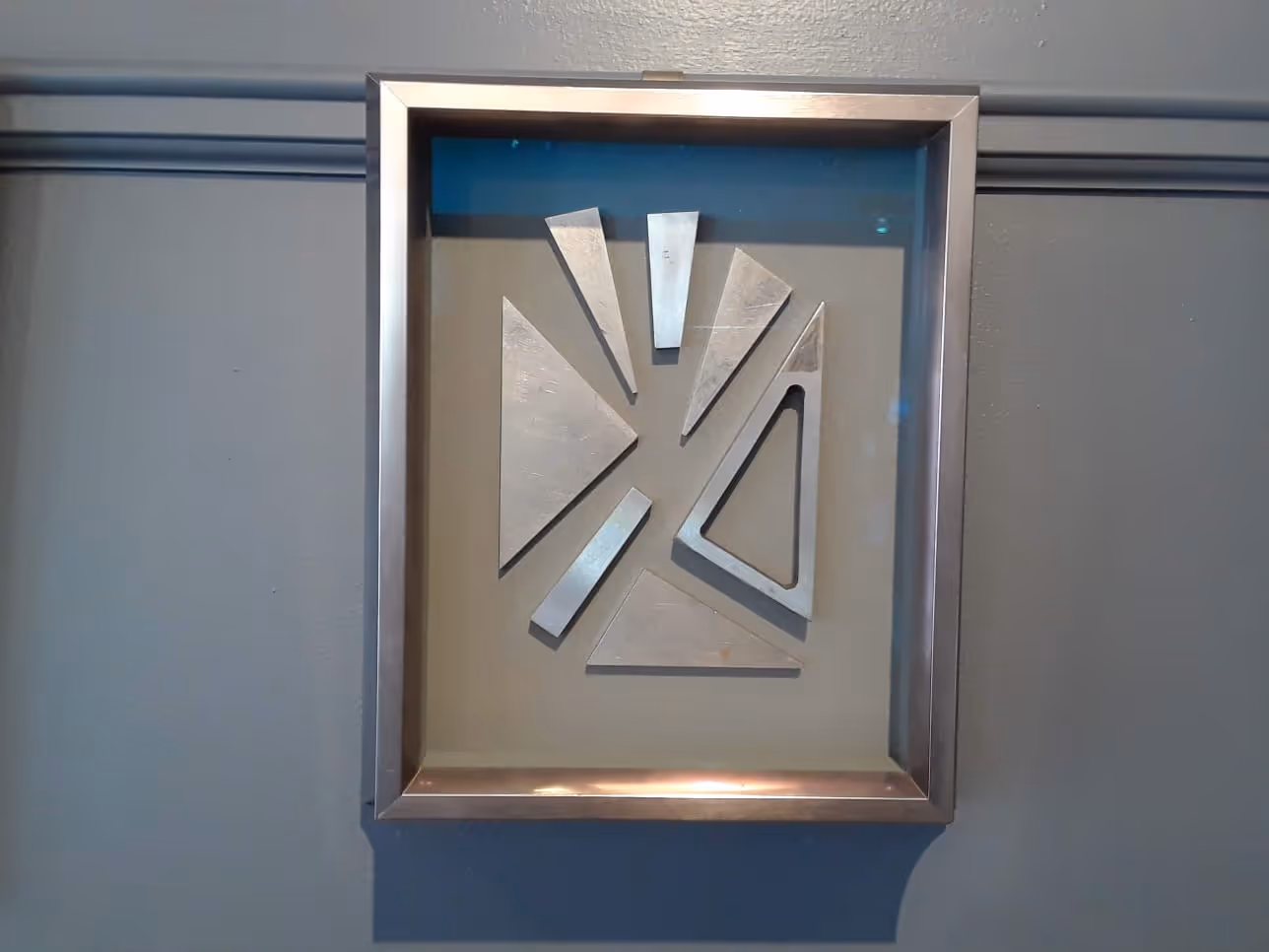 Framed abstract wall art with seven silver geometric shapes arranged on a gray background.