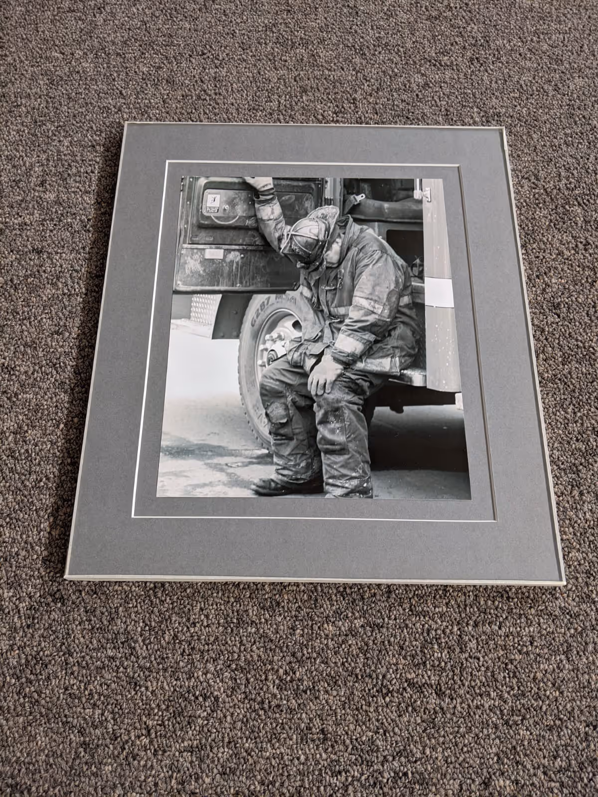 Black and white framed photograph of a firefighter sitting on the edge of a fire truck looking down, wearing full gear and helmet.