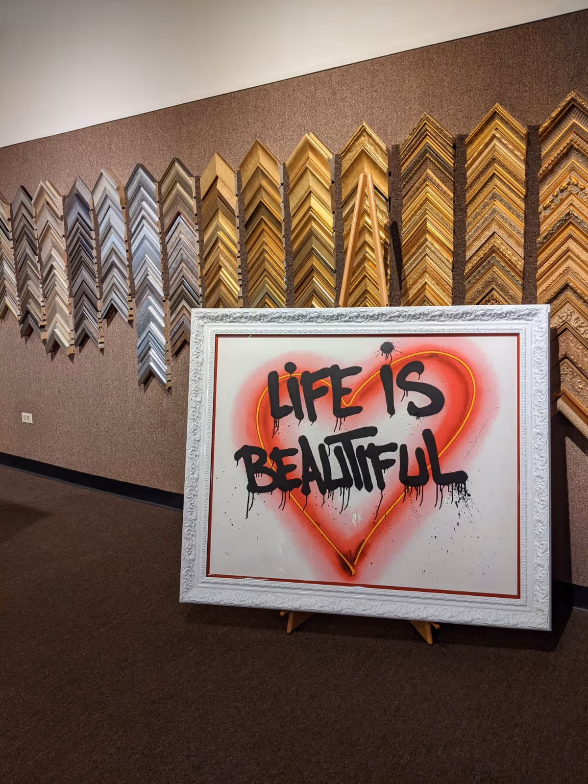 Framed artwork with a red heart and black graffiti text saying 'Life is Beautiful,' displayed in front of a wall with various ornate picture frame samples.
