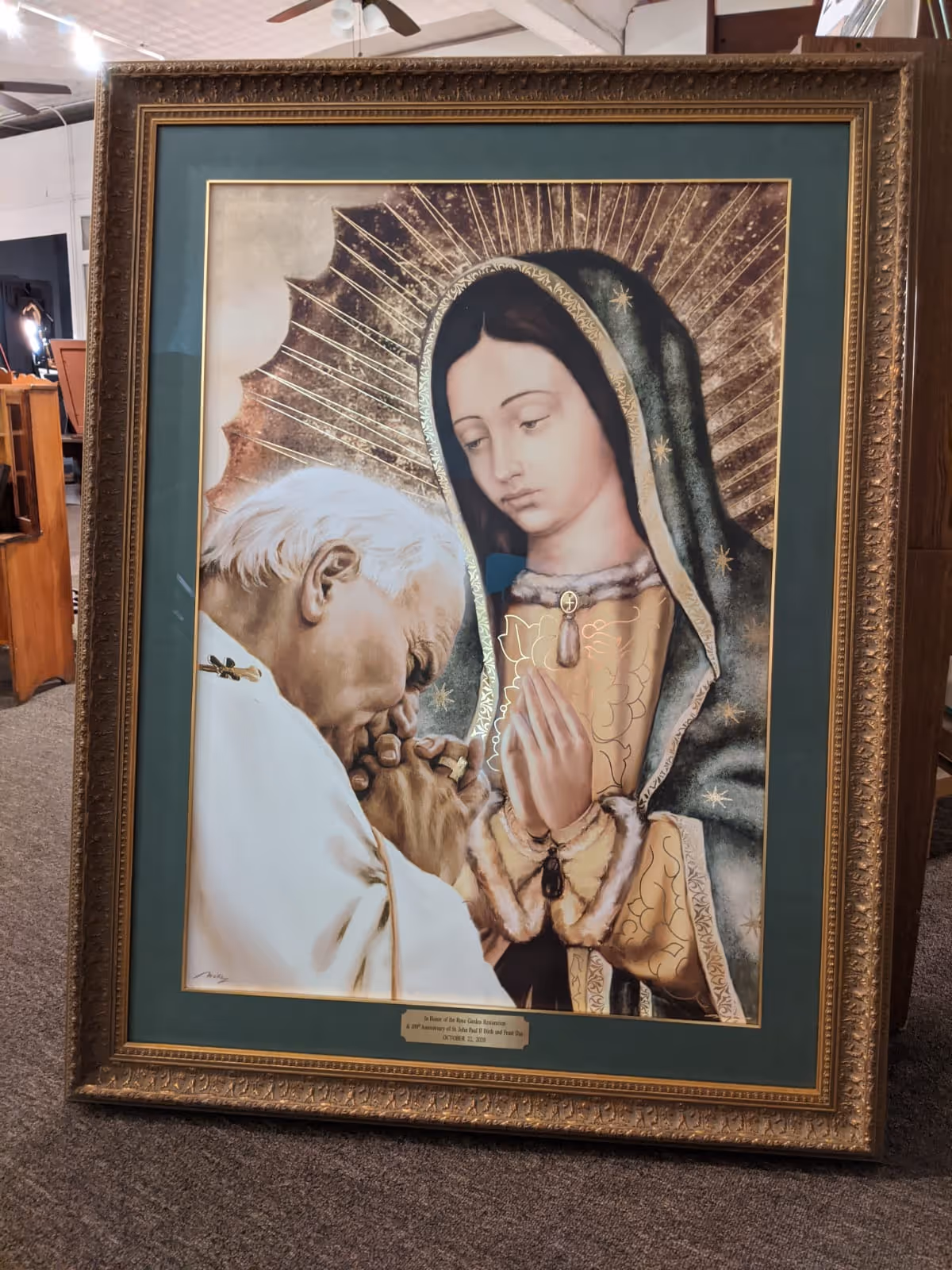 Framed religious artwork depicting a man with white hair in white robes bowing and kissing the hands of a praying woman wearing a green hooded mantle with gold rays behind her.