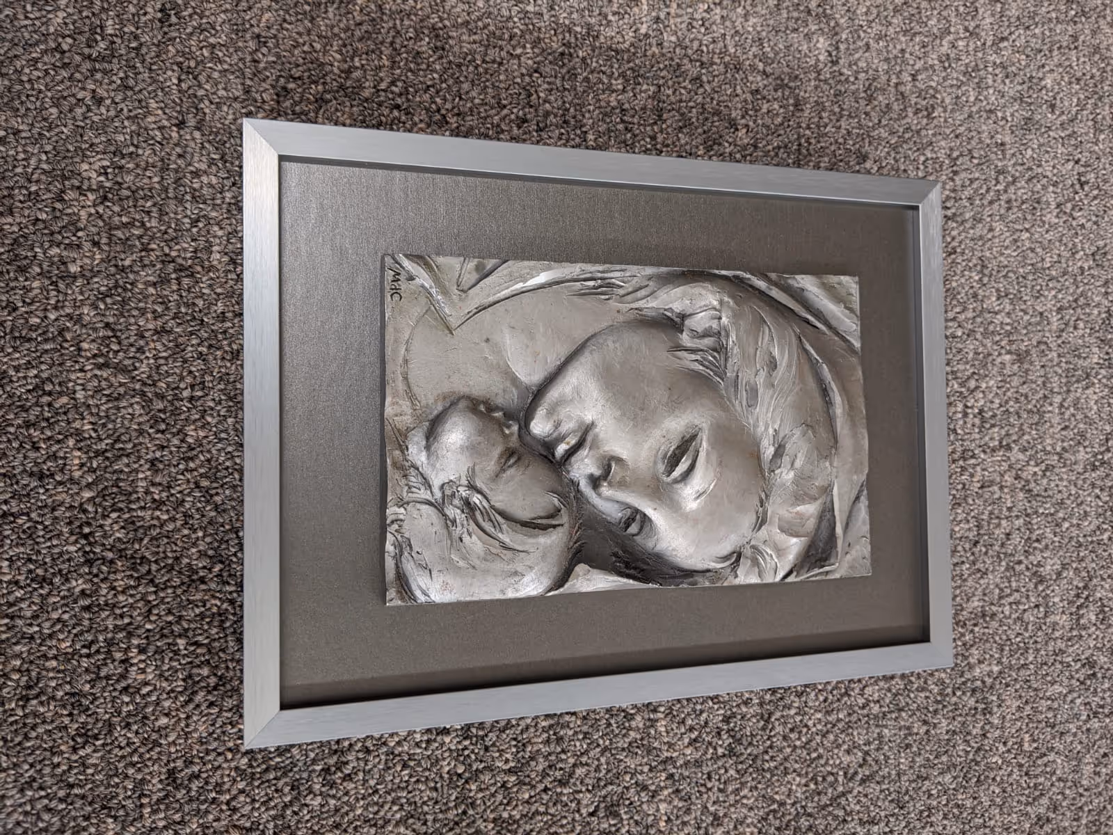 Gray framed relief artwork depicting a mother tenderly holding a child, placed on a textured carpet.