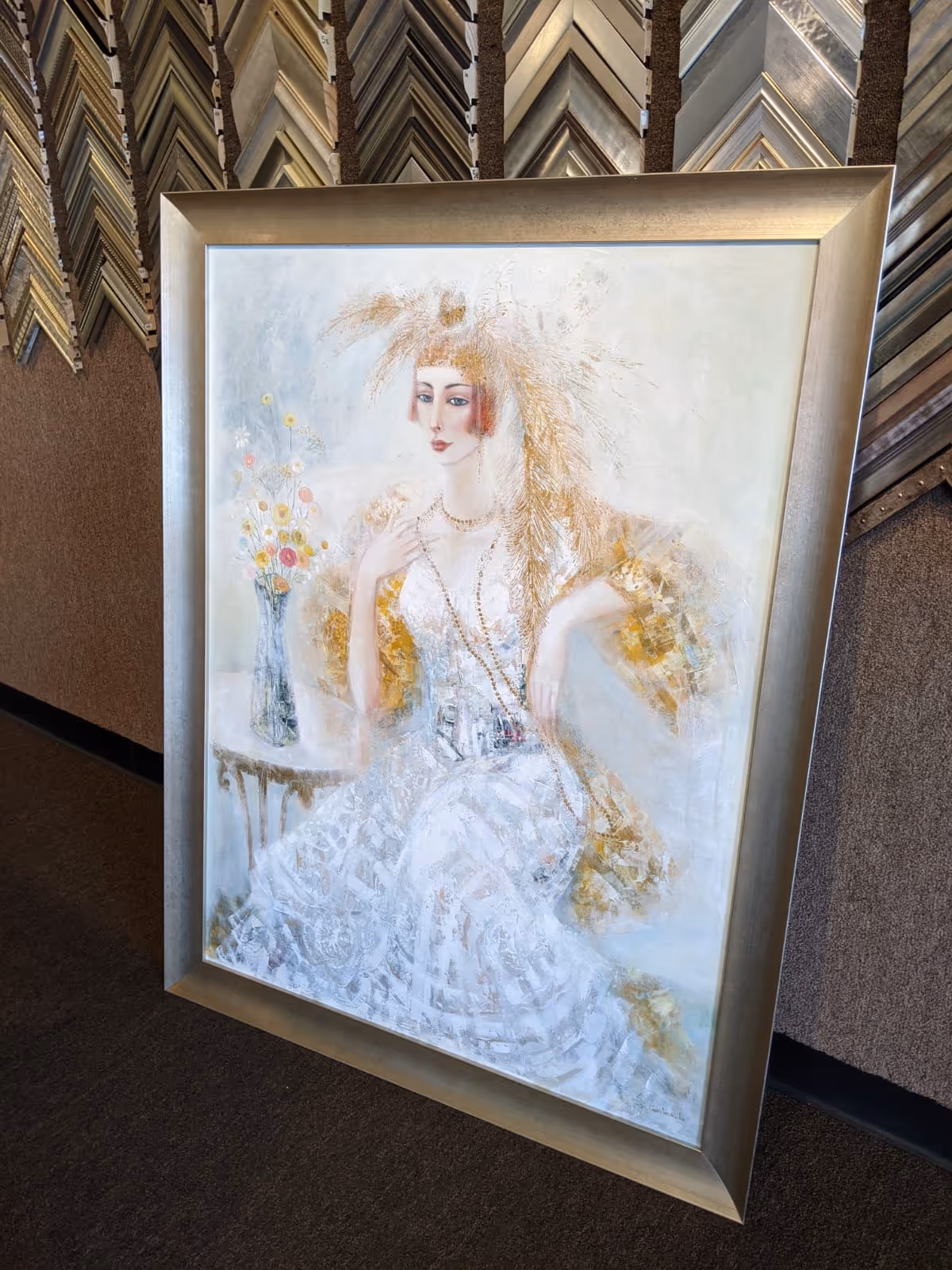 Framed painting of a woman in a white dress with an elaborate golden headdress sitting beside a table with a vase of flowers.