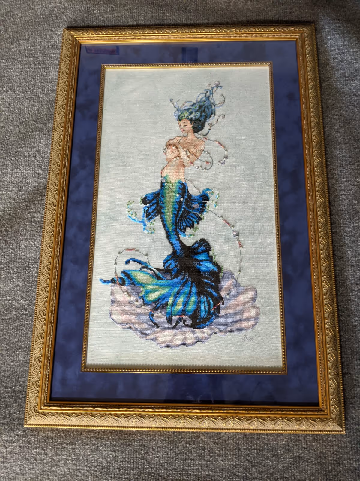 Framed embroidered artwork of a mermaid with blue and green scales sitting on a large seashell, her hair flowing with delicate swirling lines around her.