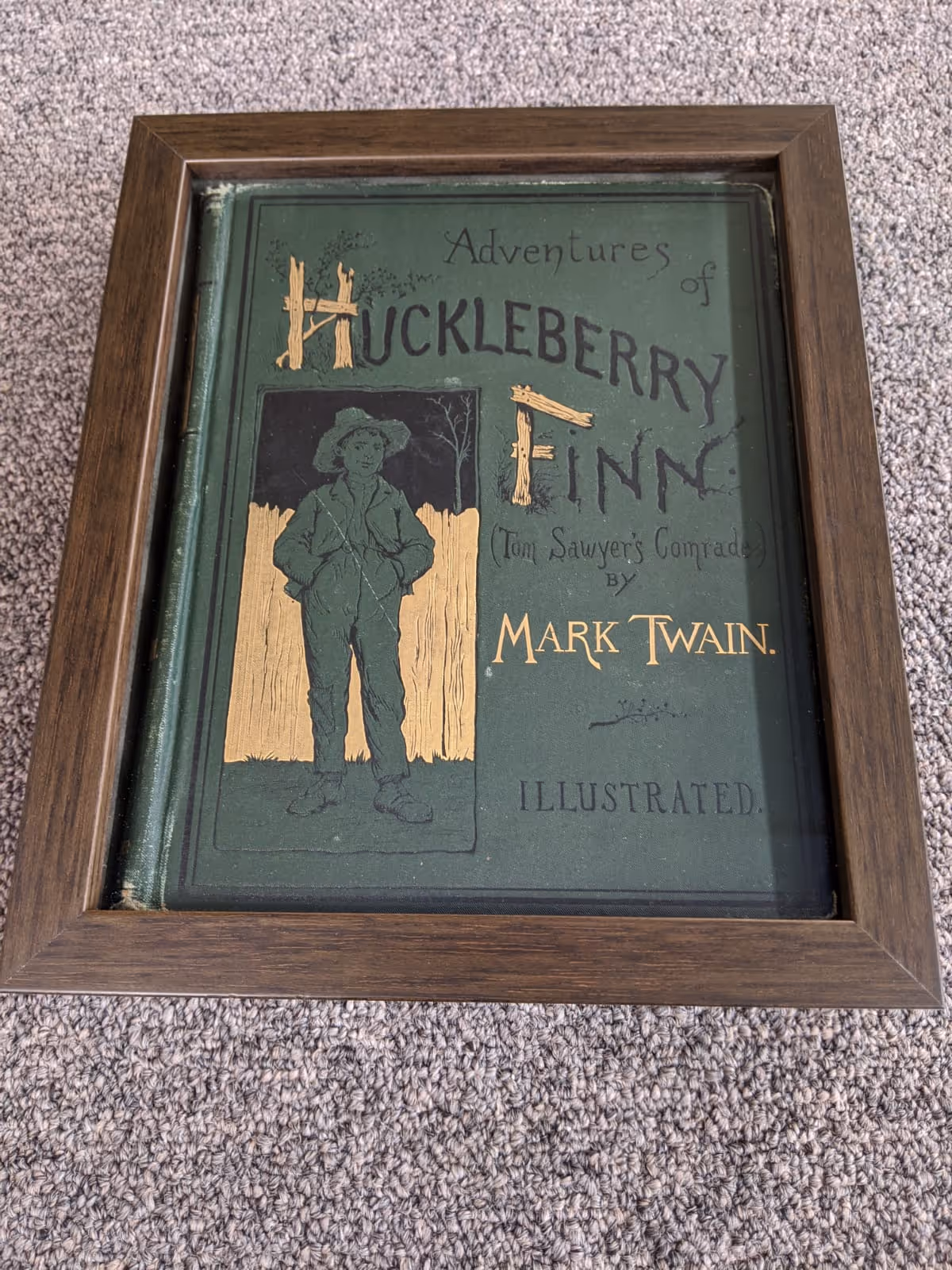Framed vintage cover of Adventures of Huckleberry Finn by Mark Twain, featuring an embossed illustration of a boy in a hat standing in front of a wooden fence.