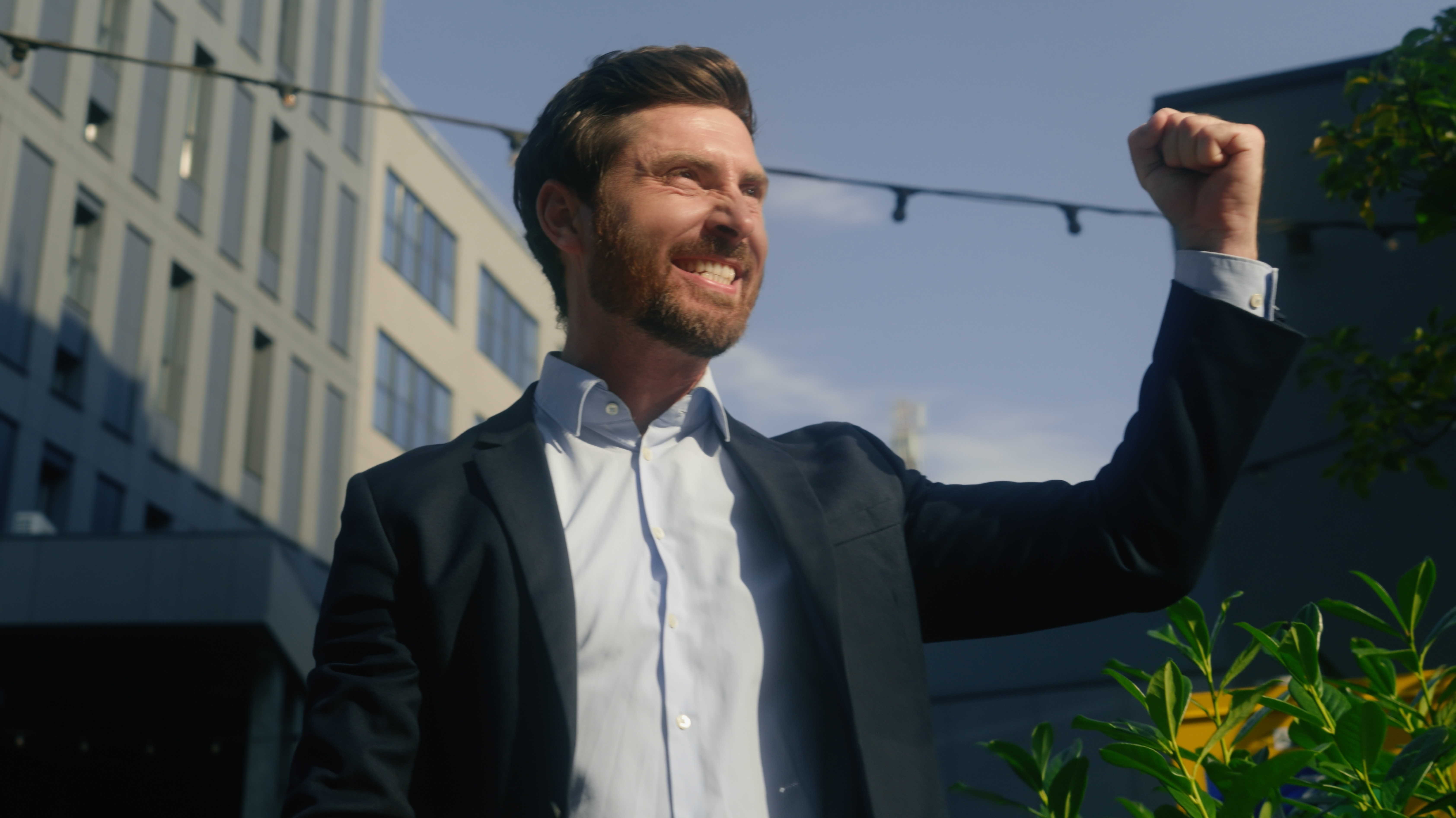 Bearded man in a suit raising his fist triumphantly outdoors near modern buildings.
