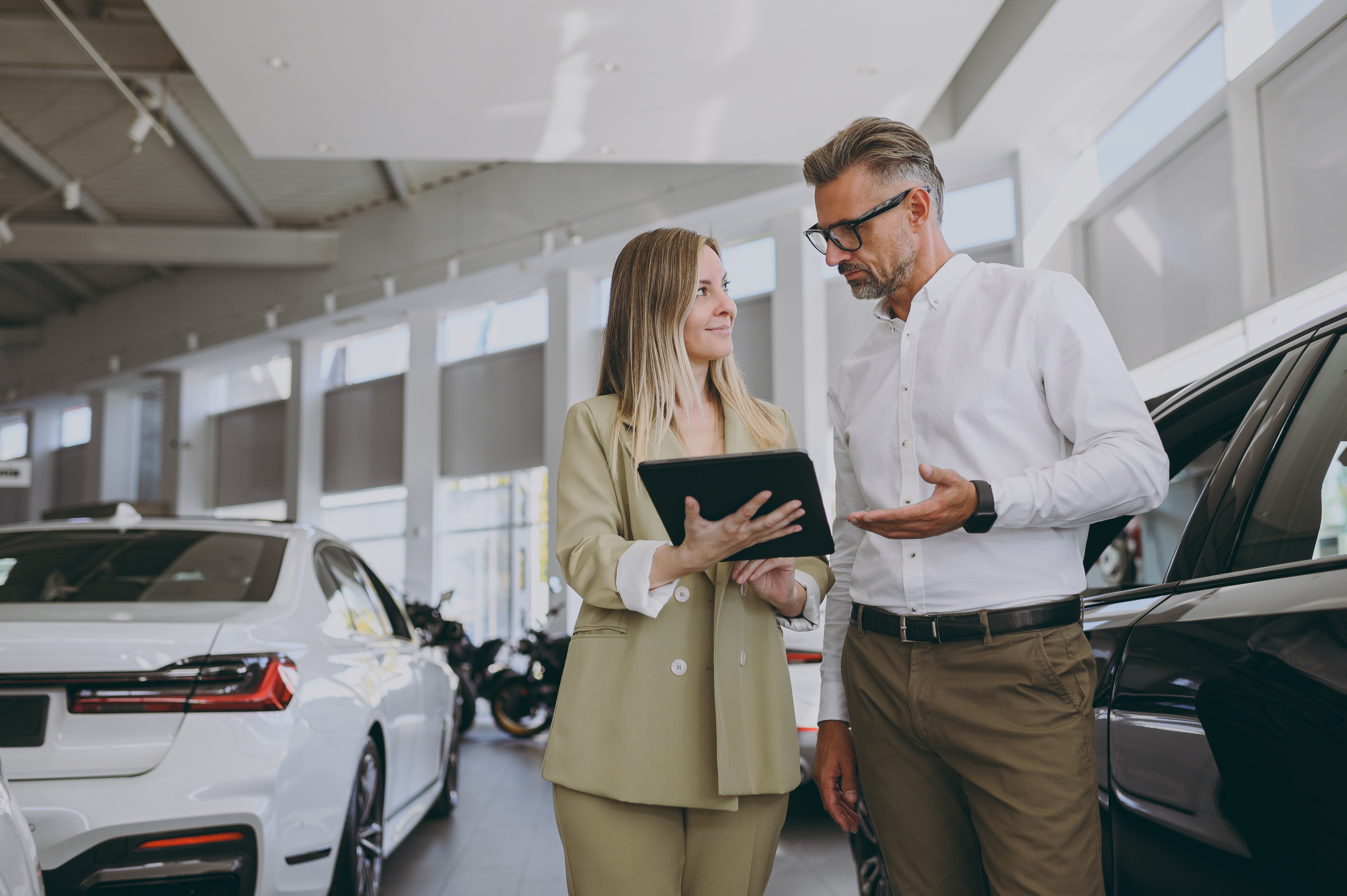 A woman in a light green suit showing a tablet to a man in glasses and a white shirt inside a car showroom.