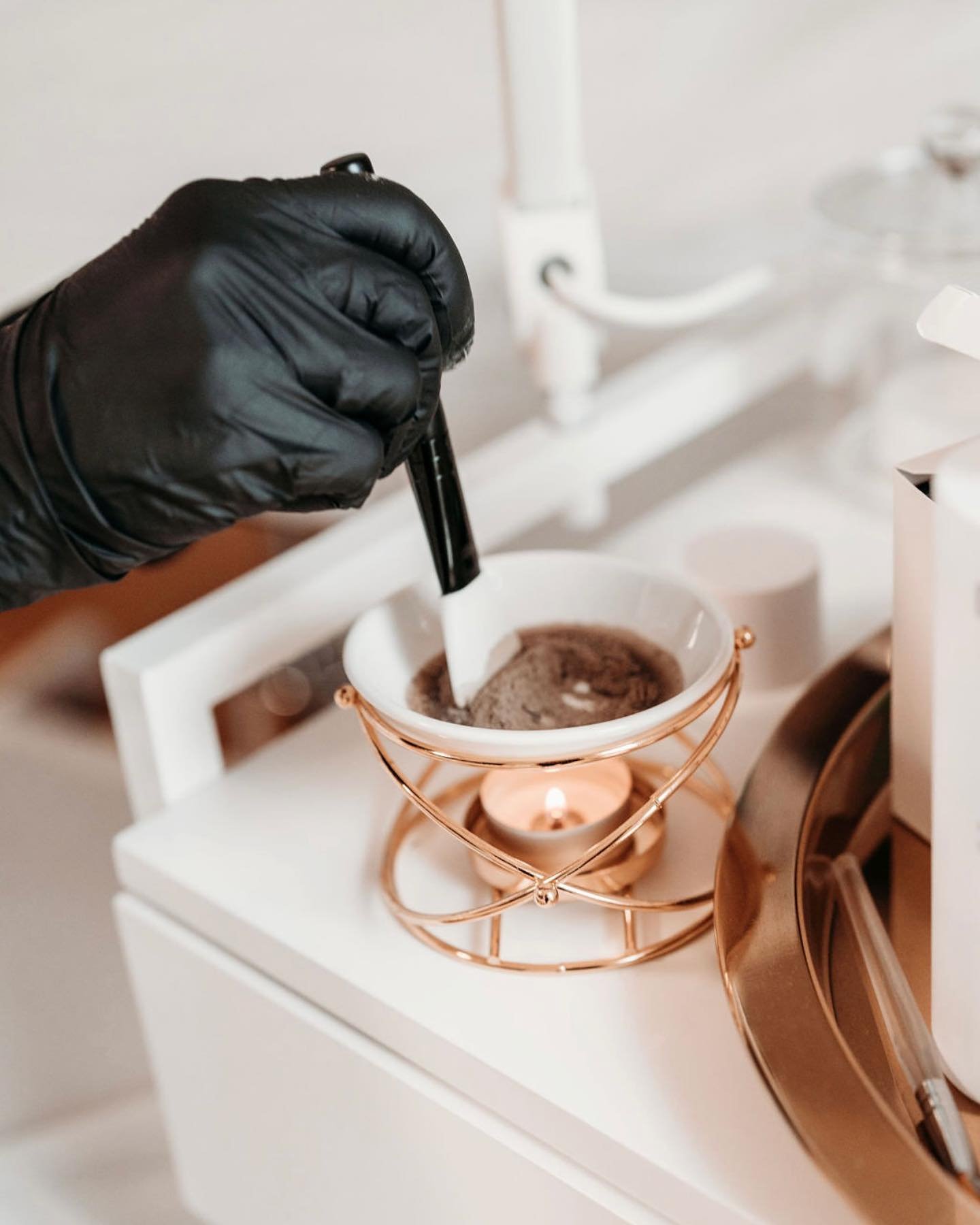 Hand in schwarzem Handschuh rührt braune Mischung in einer weissen Schale um, die von einem Teelicht in goldenem Halter erwärmt wird.