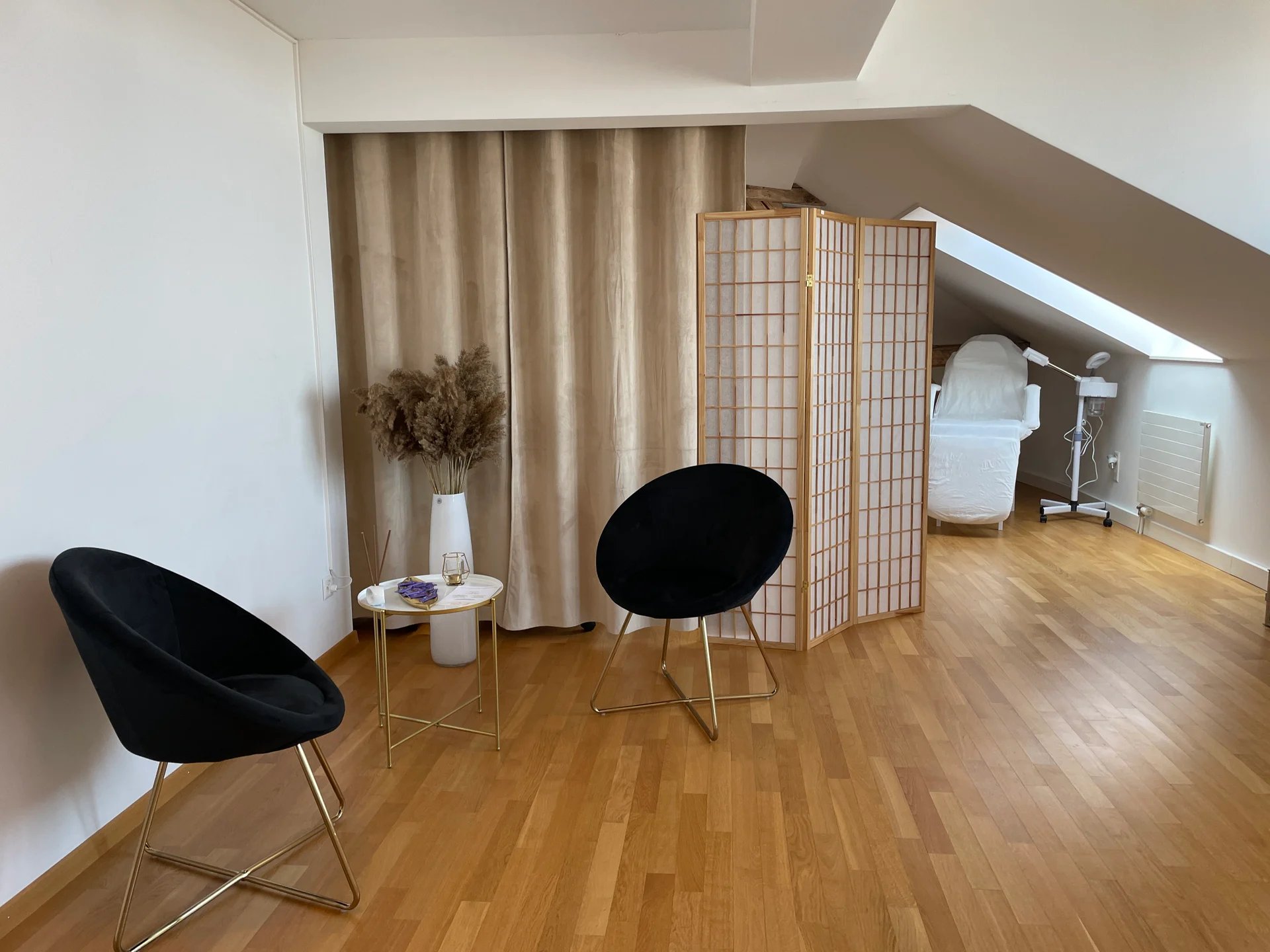 Minimalistischer Raum mit zwei schwarzen Stühlen, einem kleinen runden Tisch mit Trockenblumen und Räucherstäbchen, einem Paravent und einer weiss bezogenen Liege unter einem Oberlicht.