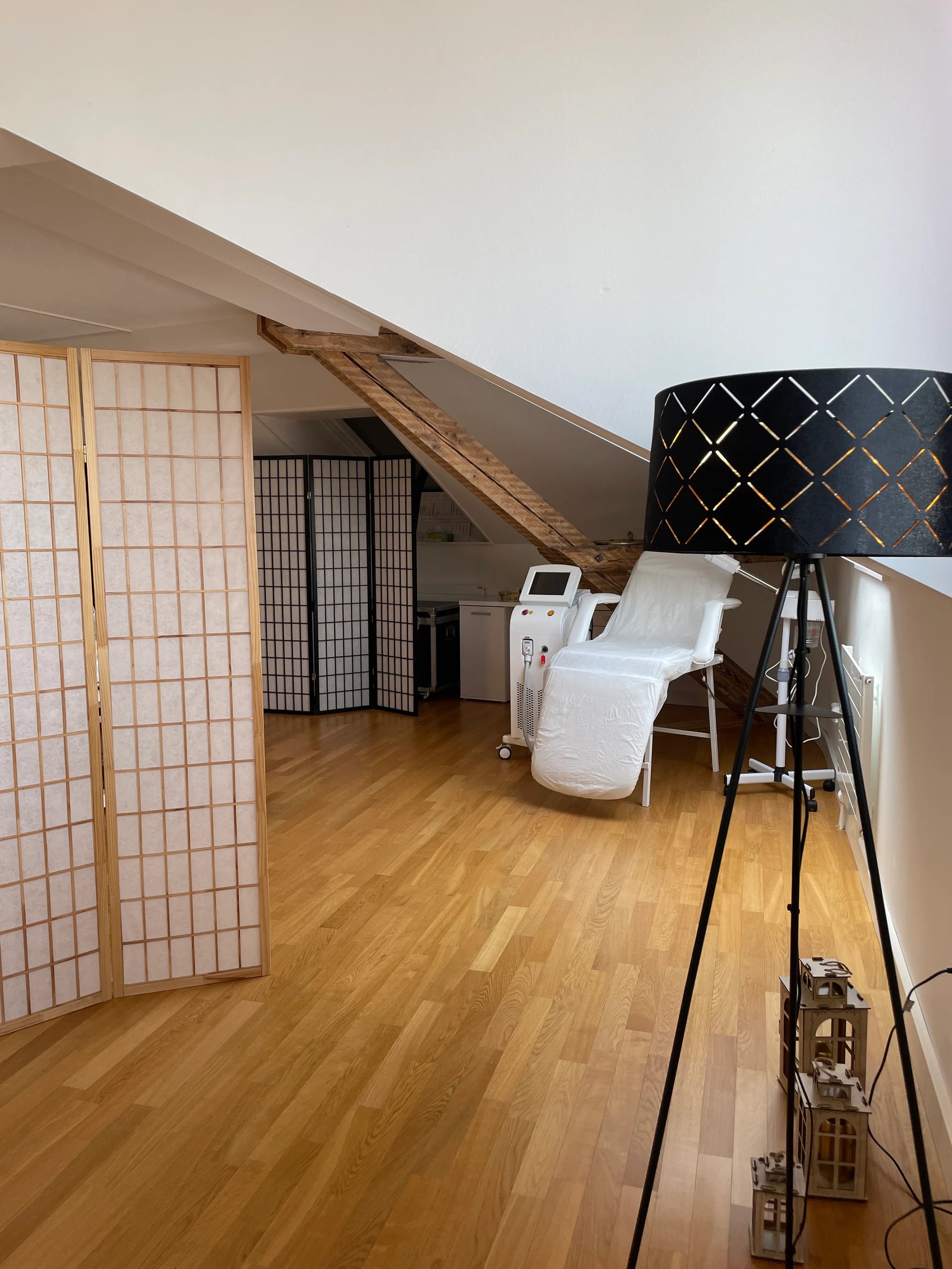 Moderner Spa-Behandlungsraum mit Holzboden, weisser Liegestuhl, Sichtschutzwänden und Stativlampe mit schwarzem Schirm.