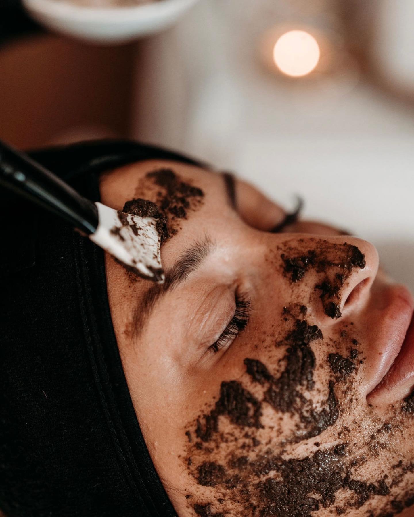 Nahaufnahme einer Person mit geschlossenen Augen, die eine dunkle Gesichts-Peeling-Maske erhält, die mit einem Spatel aufgetragen wird.