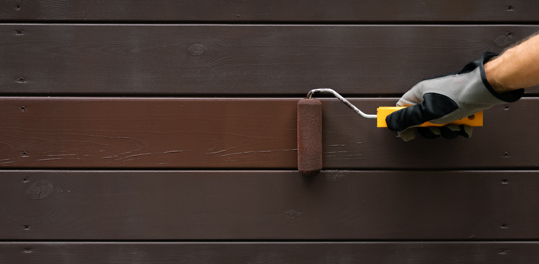 Hånd med hanske maler utendørs trepanel med brun maling og malerull.
