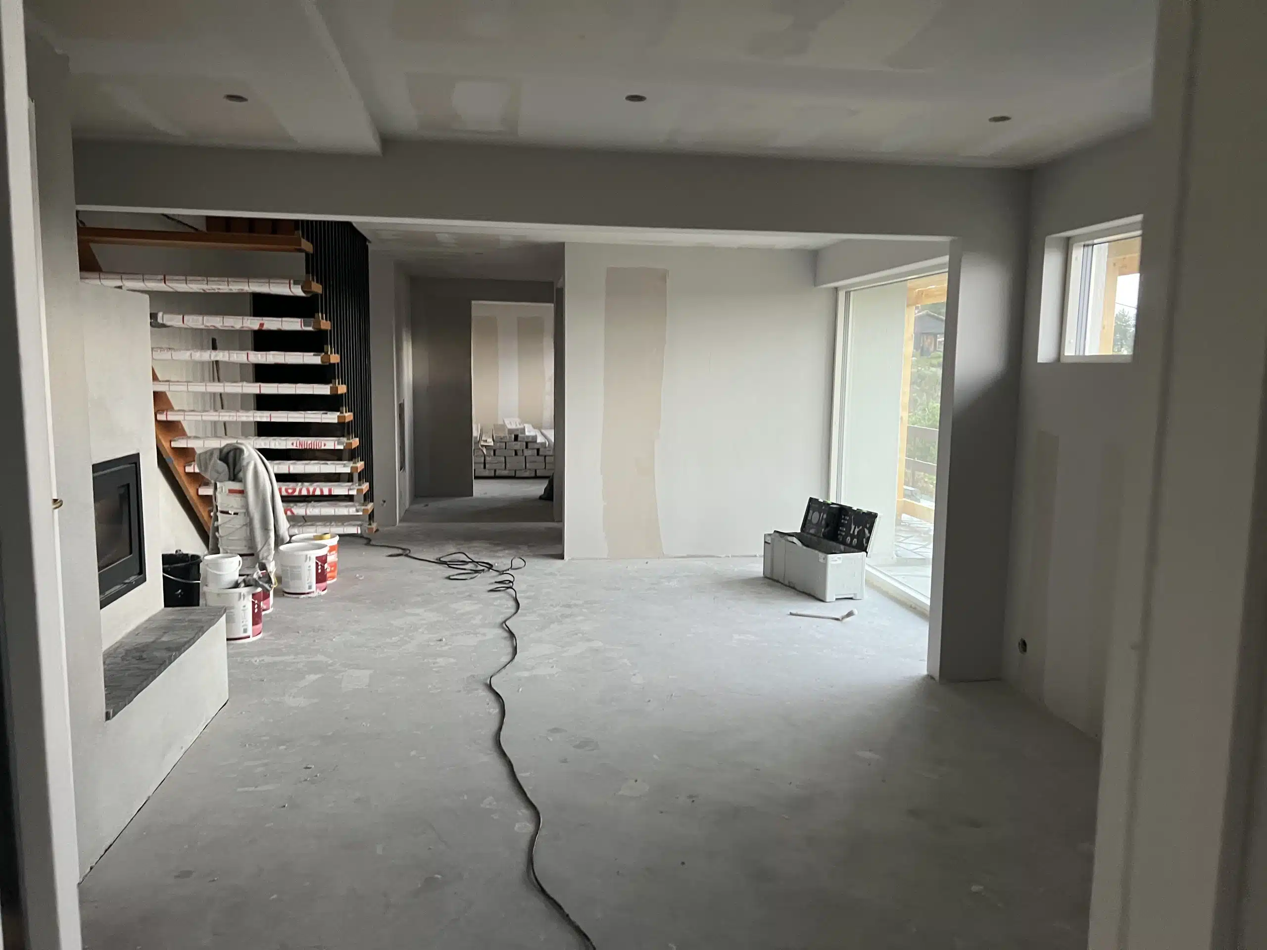 Rom under oppussing med trapp dekket med beskyttelse, malingsspann og verktøykasse på betonggulv.