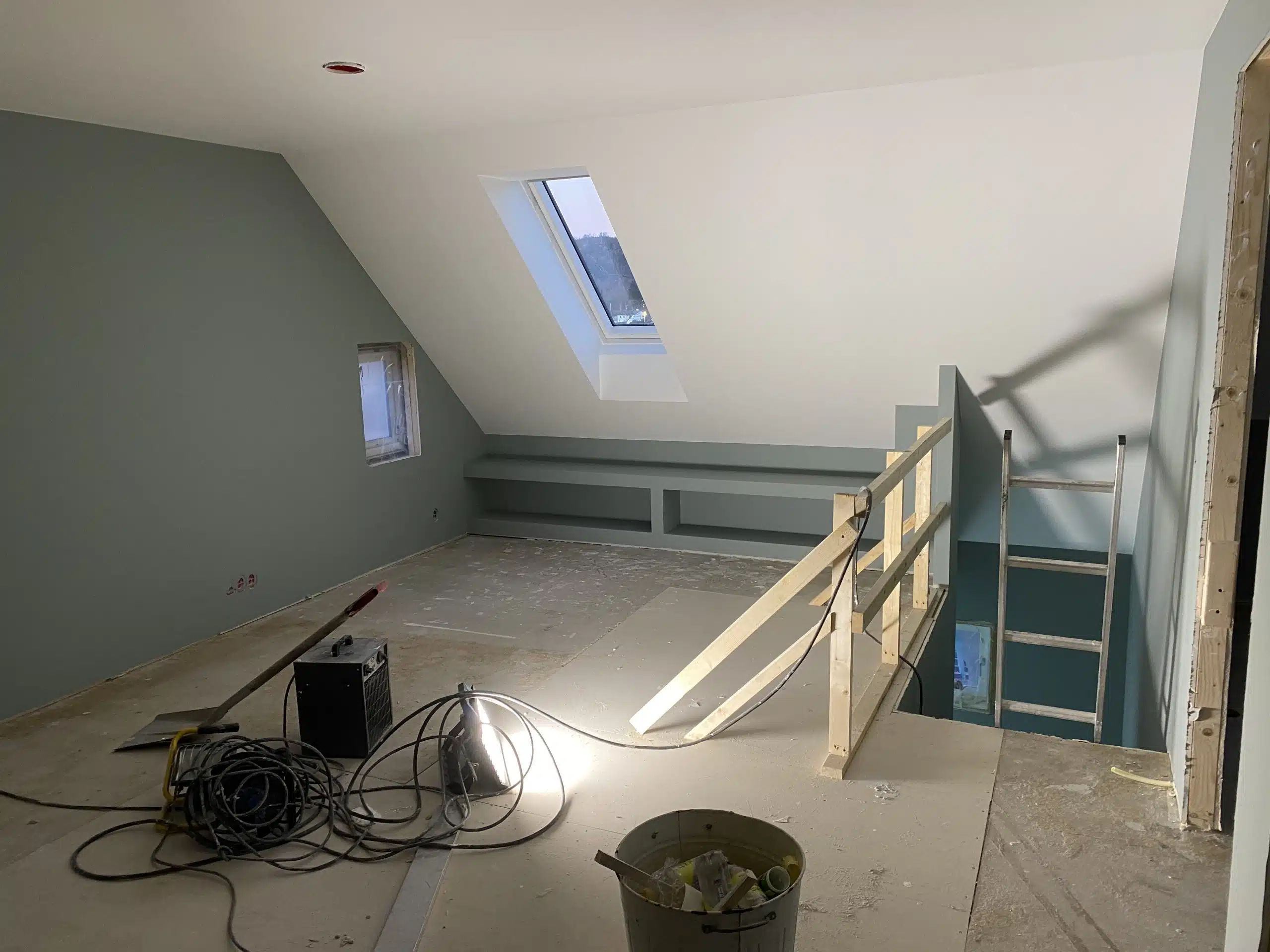 Mellomrom under oppussing med skråtak, et vindu i taket, konstruksjonsmaterialer, og en stige ved trappeåpningen.