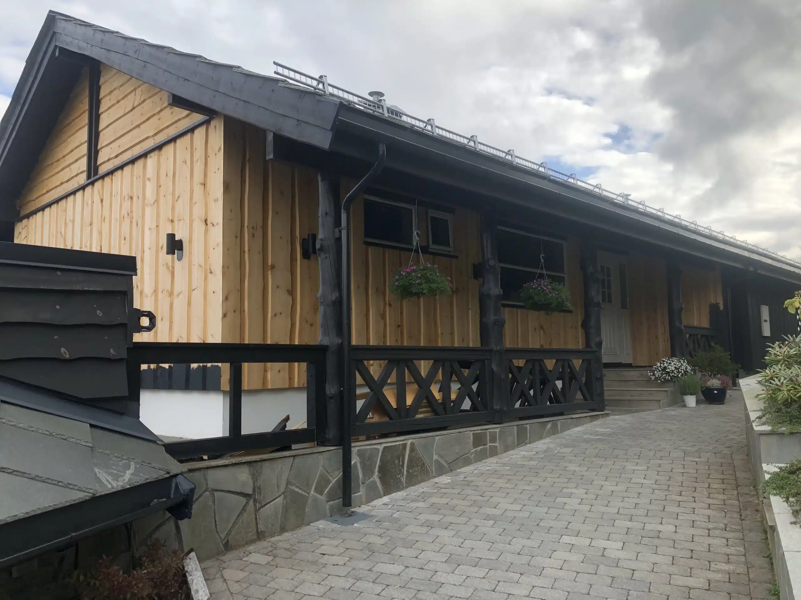 Trehus med lys trepanel, svart tømmer, blomsterpotter og brosteinsoppkjørsel under en skyet himmel.