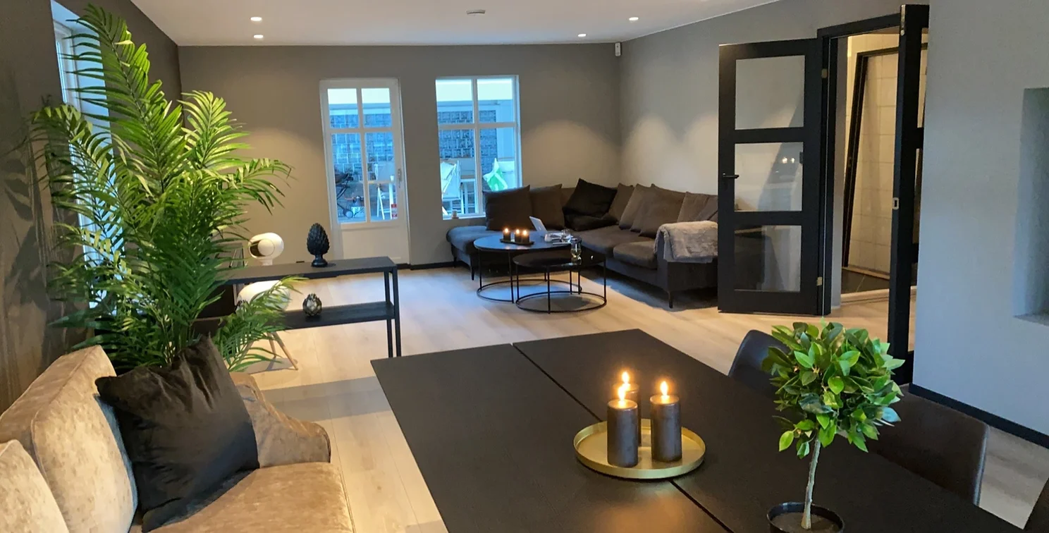 Moderne stue med beige sofa, store grønne planter, svarte bord med tente lys og en mørk hjørnesofa ved vinduene.