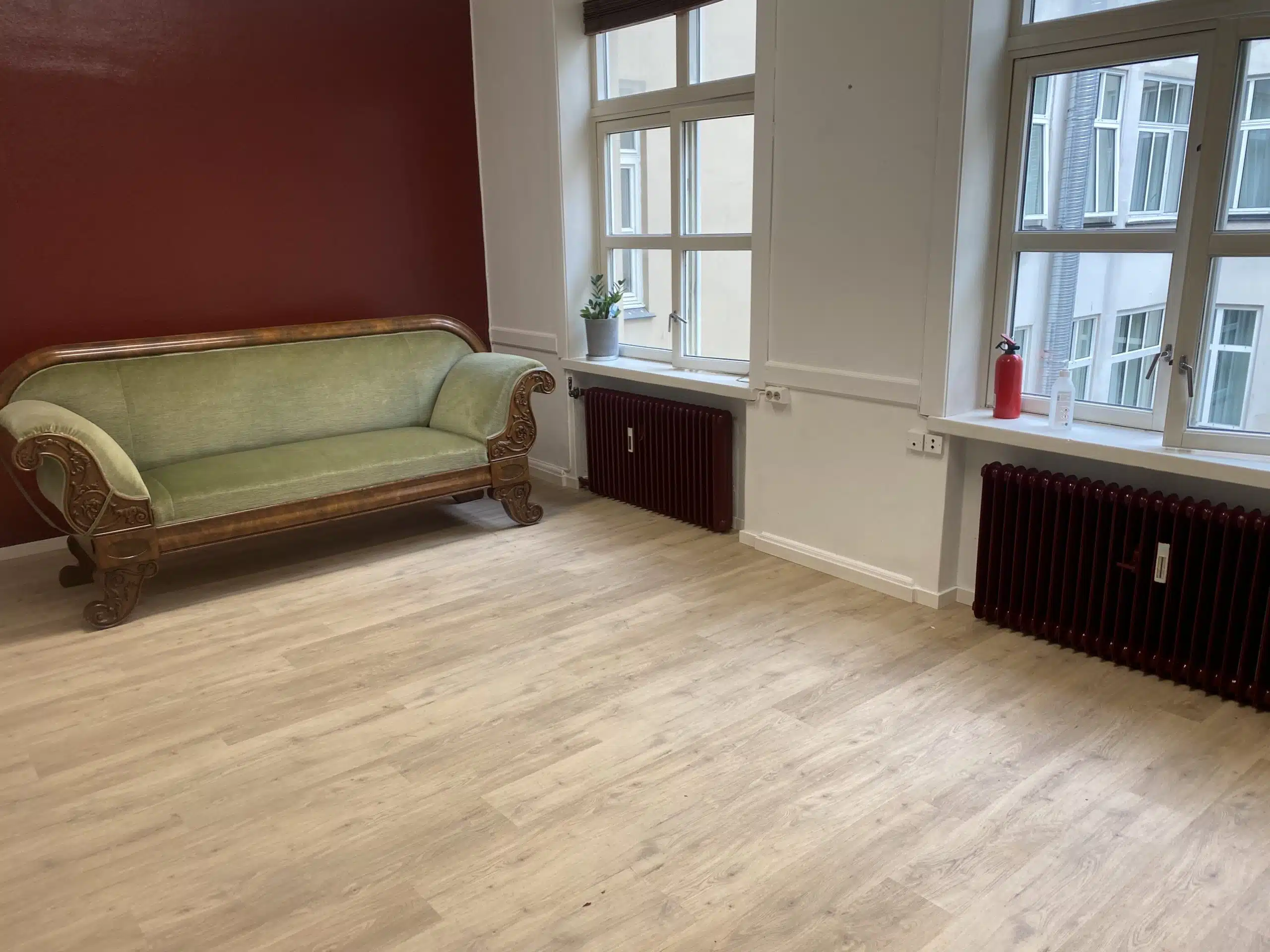 Rom med lys trefarget gulv, en antikk grønn sofa med utskåret treramme mot en mørkerød vegg, to store vinduer med hvite karmer, to mørkerøde radiatorer, en brannslukker, og en plante på vinduskarmen.