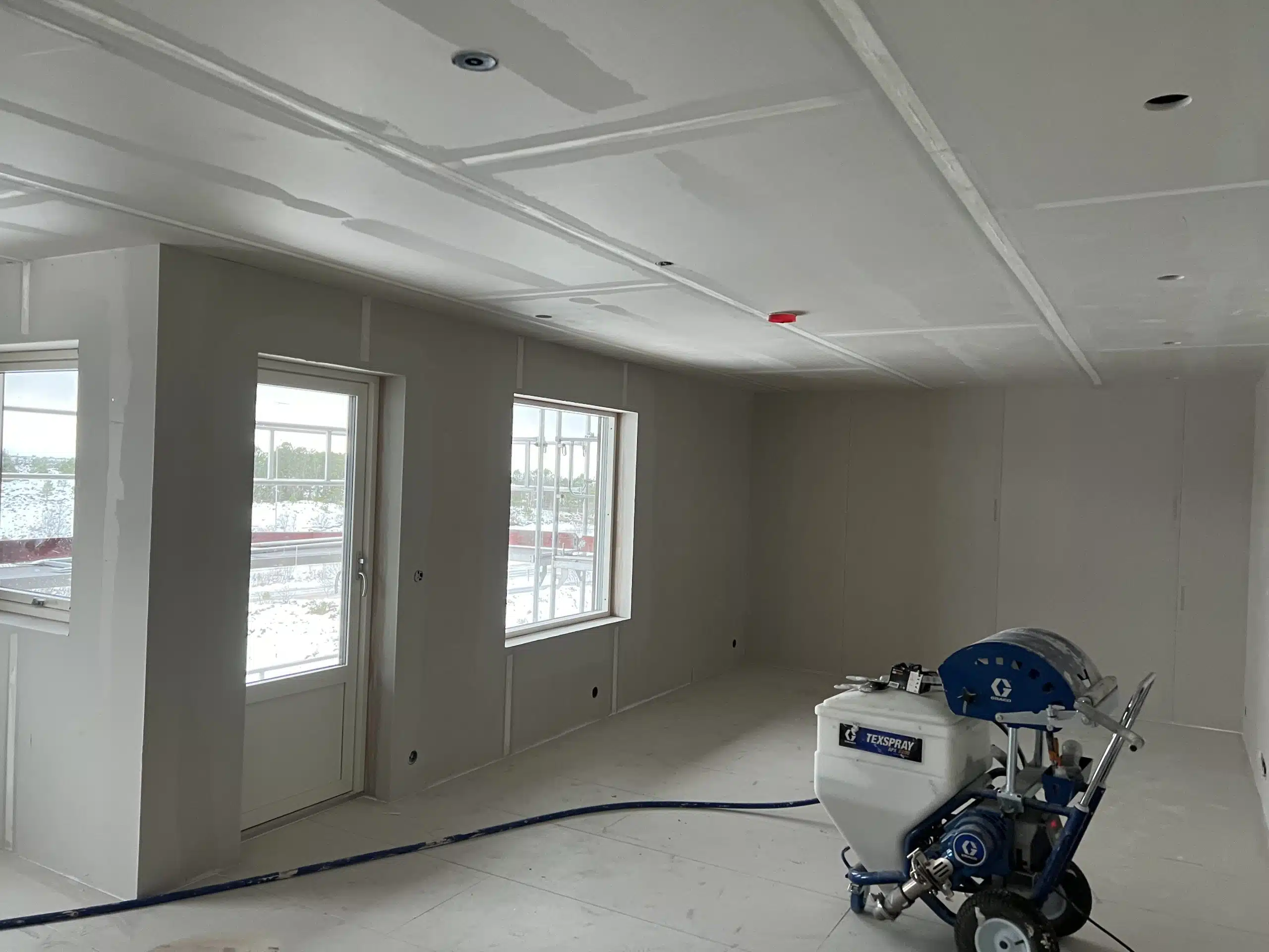 Et rom under oppussing med uferdige gipsvegger og tak, et vindu og en dør med utsikt til snødekt landskap, og en sprøytemaskin for maling på gulvet.