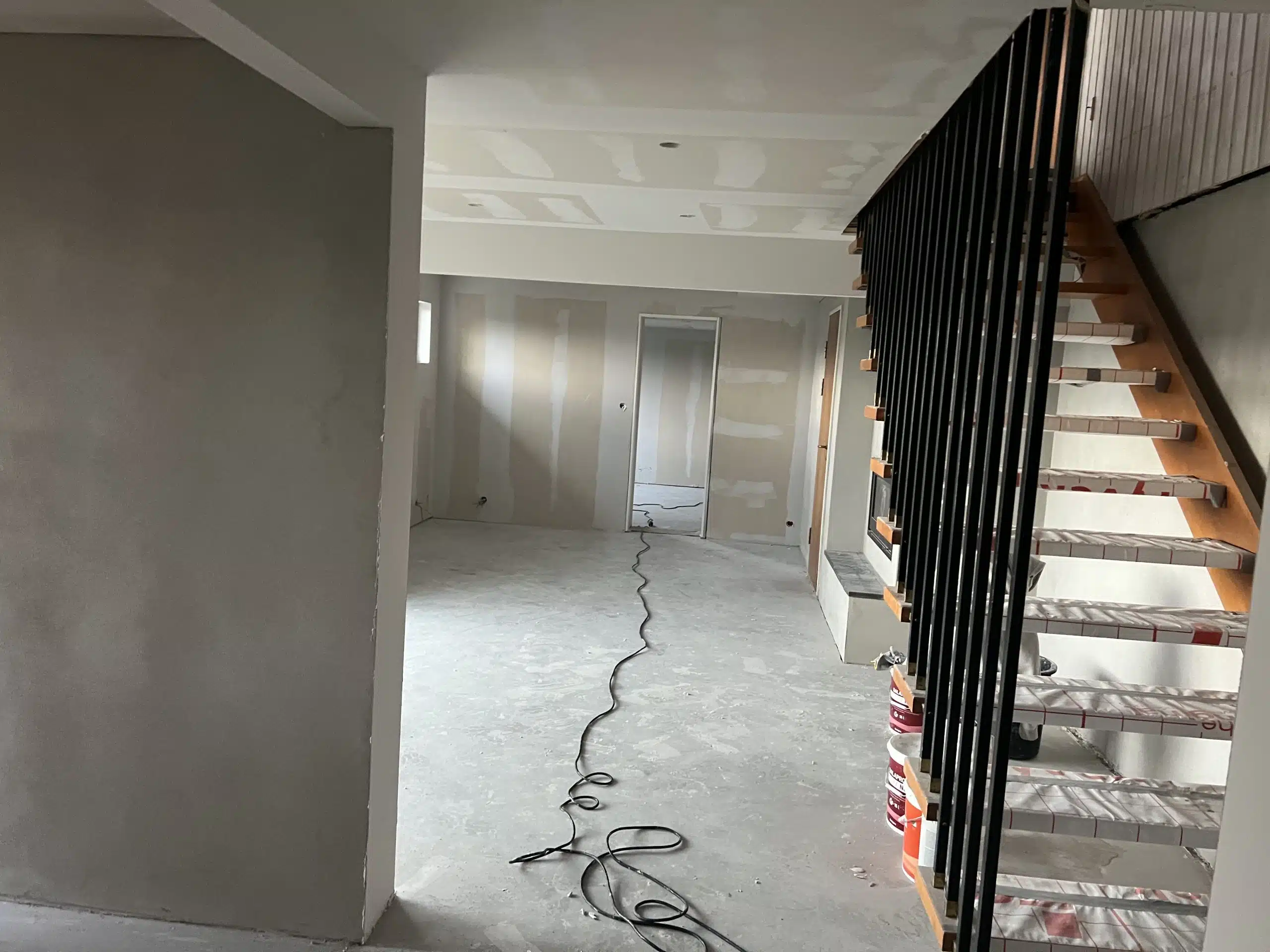 Innendørs byggeplass med uferdig vegger, en trapp med svart rekkverk til høyre og en elektrisk ledning på gulvet.