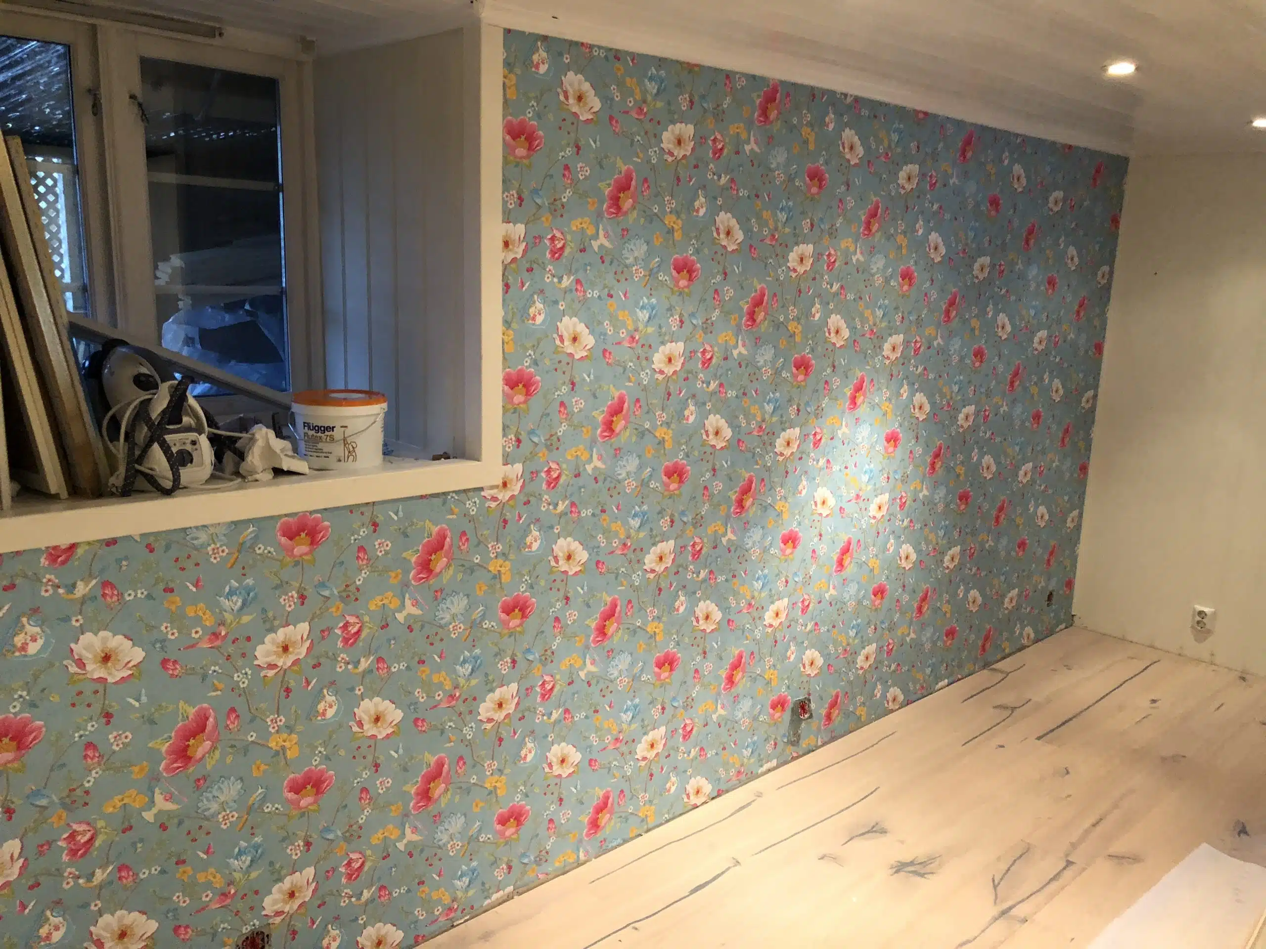 Rom med blå blomstret tapetvegg, lyst tregulv, vindu med malingsutstyr og stæsj.