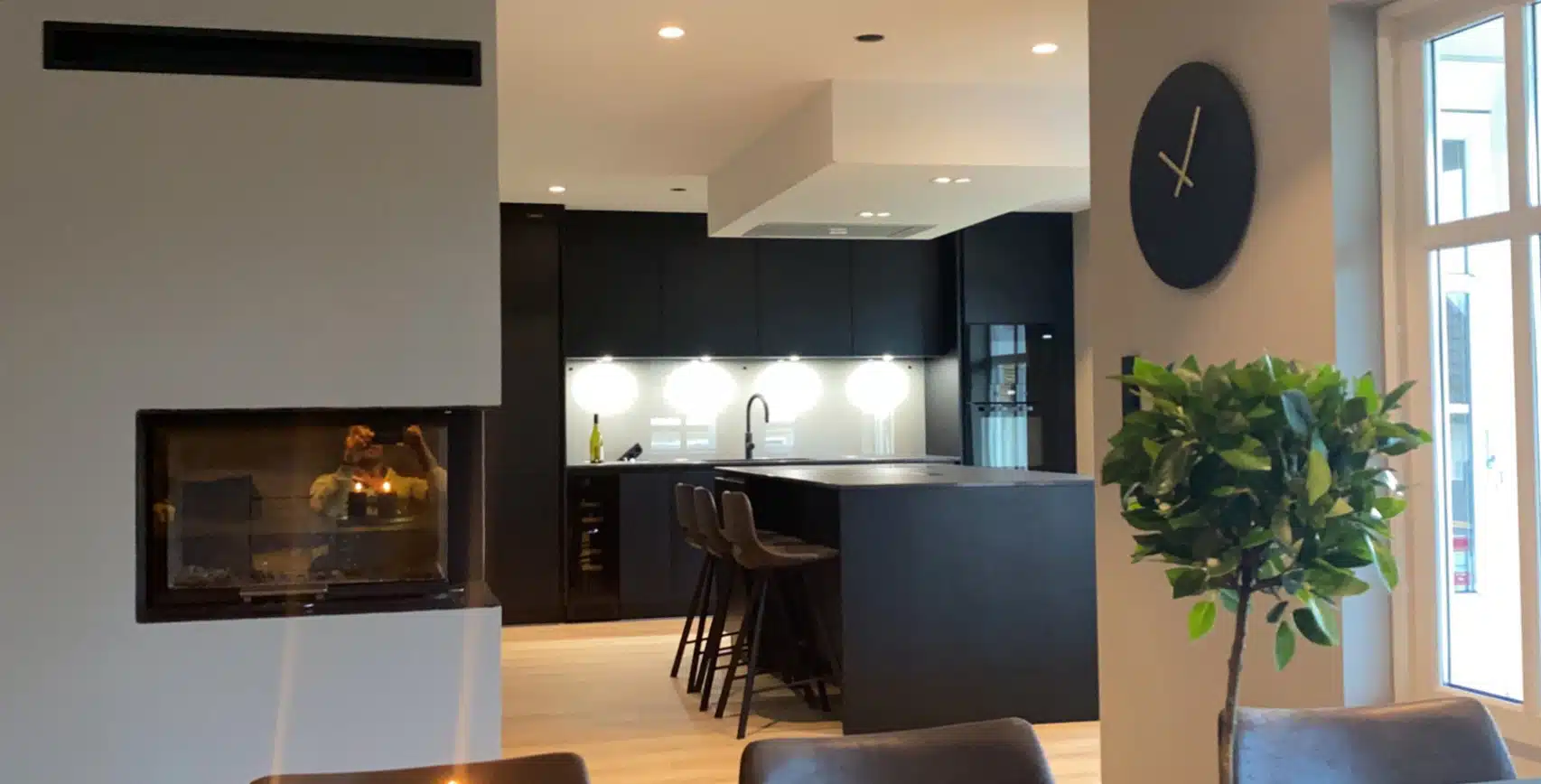 Moderne kjøkken med svart kjøkkenøy, barstoler, peis, veggur og et grønt potteplante ved vinduet.