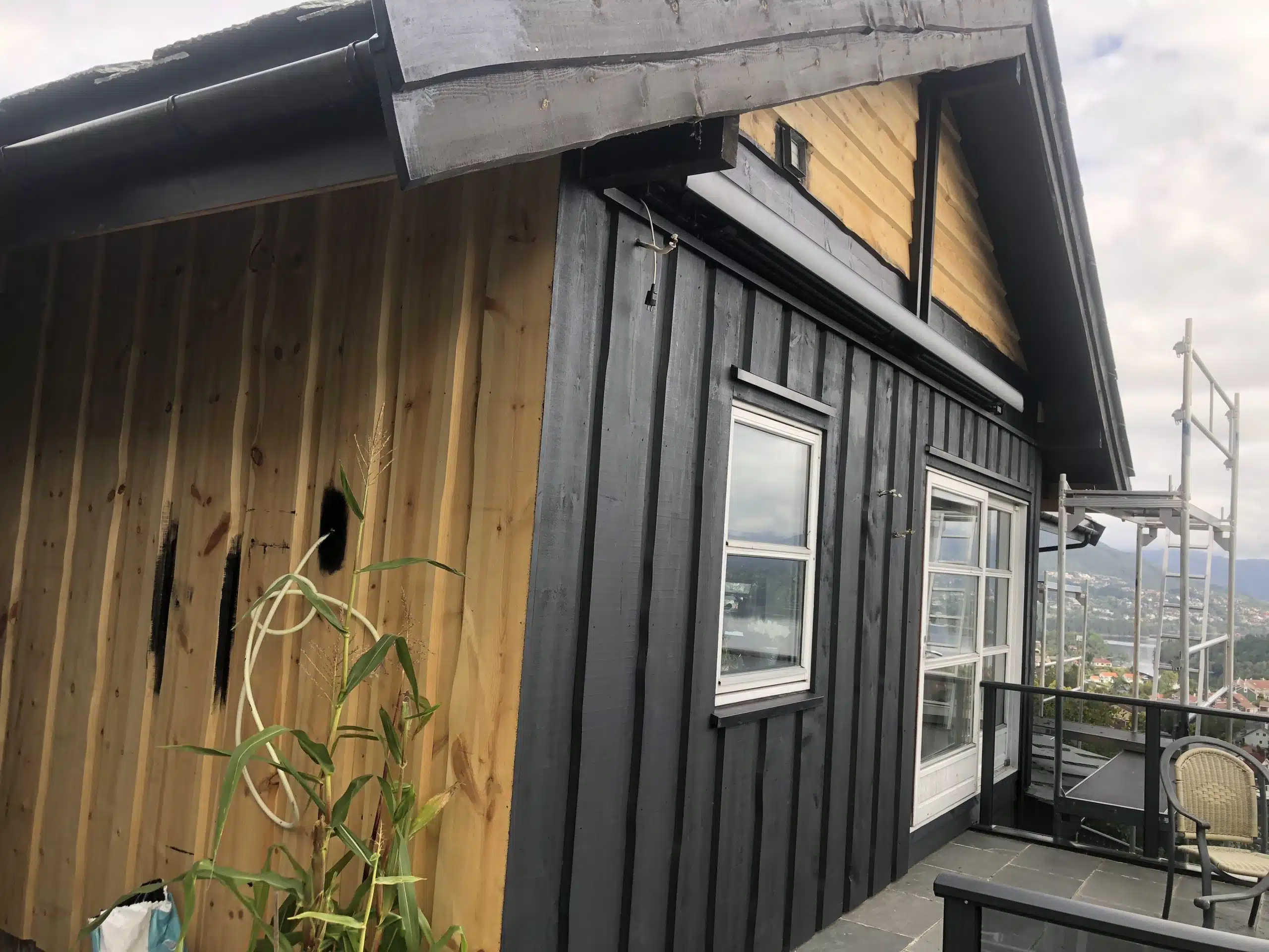 Del av et hus under oppussing med svart og ubehandlet trepanel, en balkong med glassrekkverk og utsikt over et boligområde og fjell i bakgrunnen.