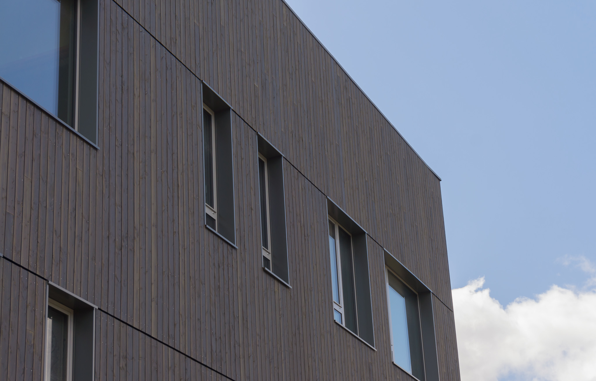 Moderne bygning med vertikale trepaneler og flere rektangulære vinduer under en blå himmel med noen skyer.